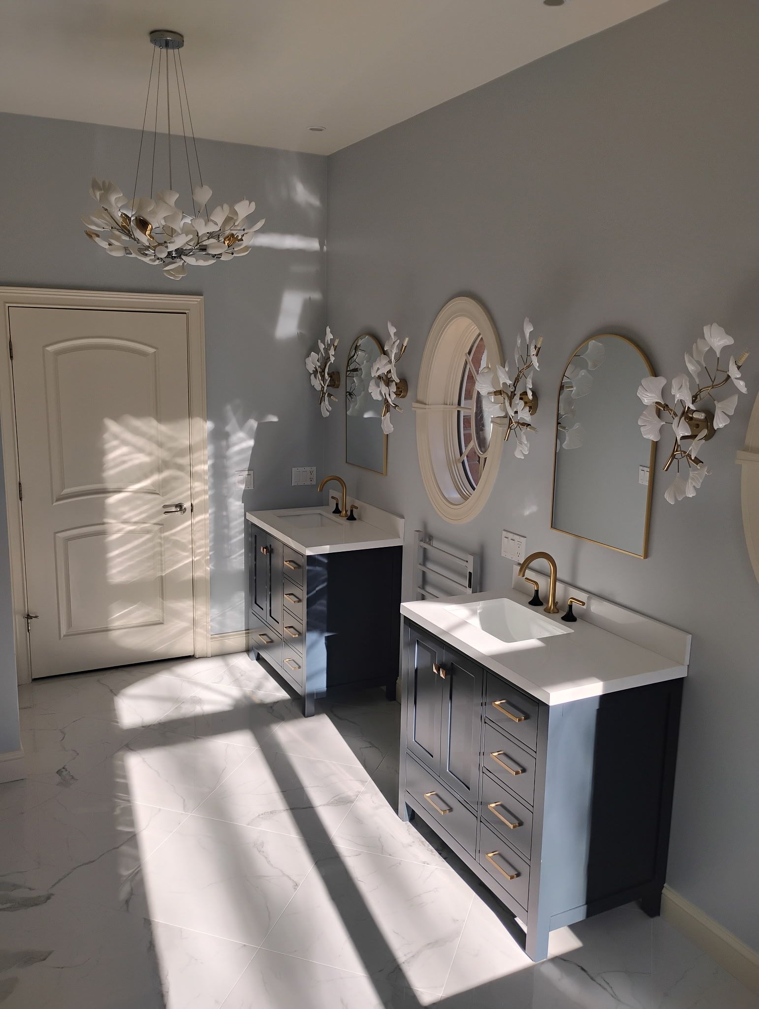 Bright white bedroom with mirrored dressers, ornate wall mirrors, chandelier, and sunlight on gray walls and floor.