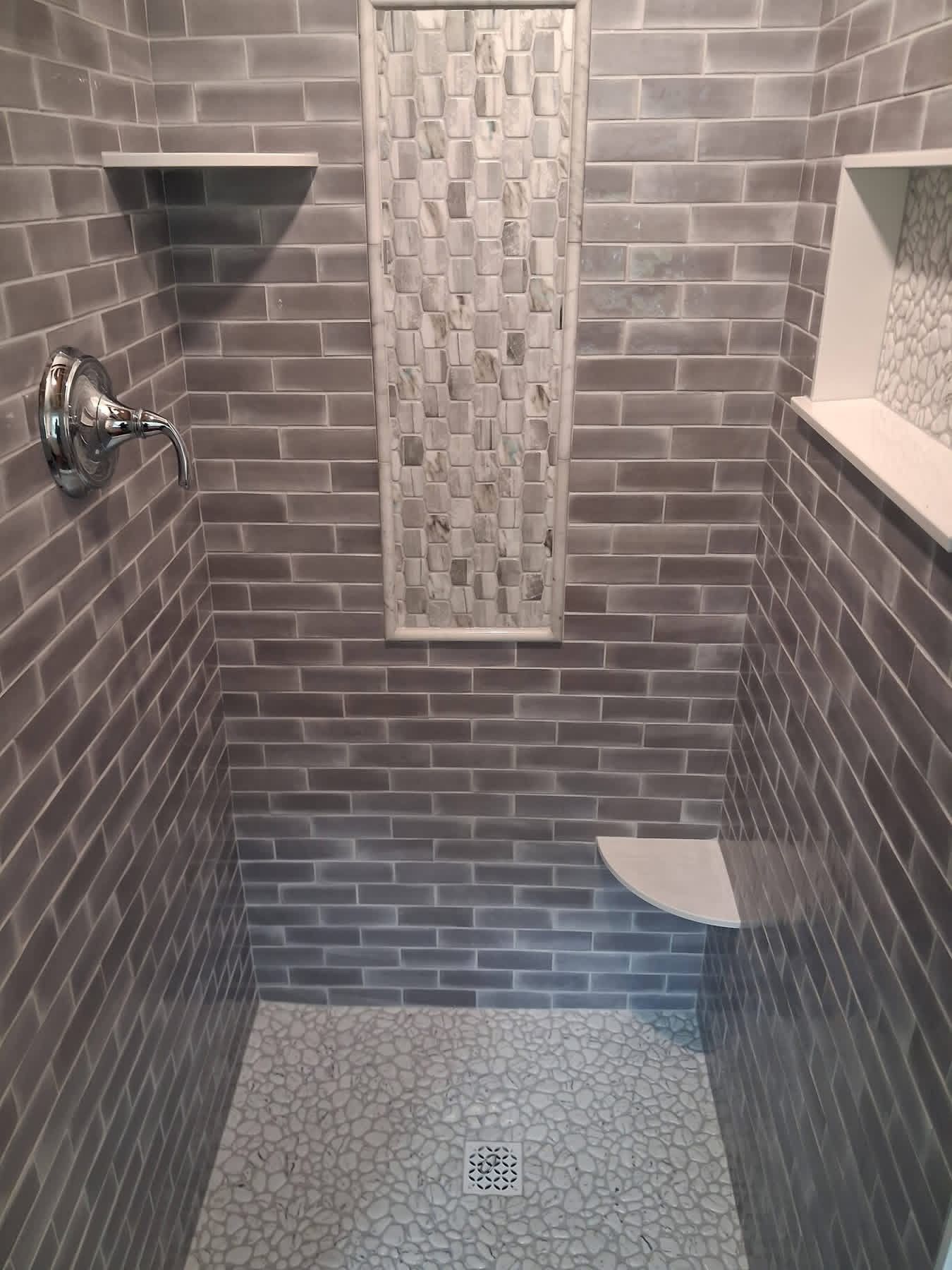 Small tiled walk-in shower with bench, recessed niche, chrome fixtures, and a narrow mosaic accent panel.