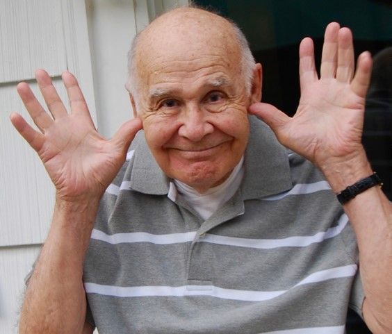 An older man wearing a grey and white striped shirt is waving his hands