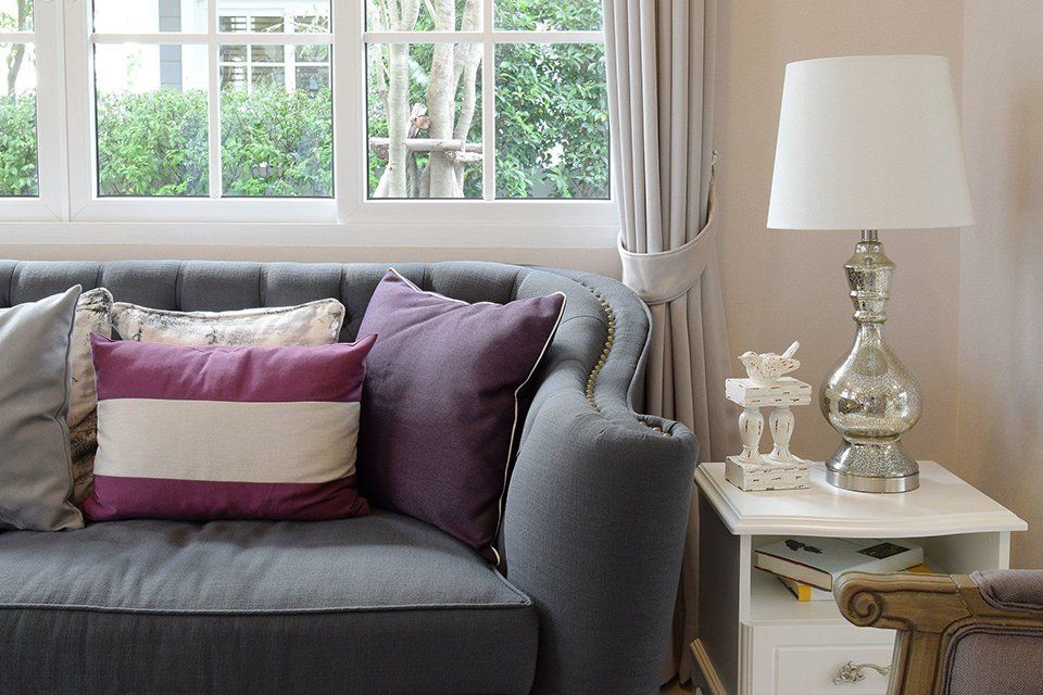 A living room with a gray couch , purple pillows , a lamp and a window.