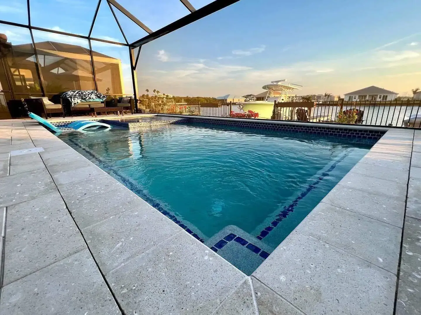 Outdoor swimming pool with clear blue water, stone deck, and covered patio overlooking a waterfront at sunset