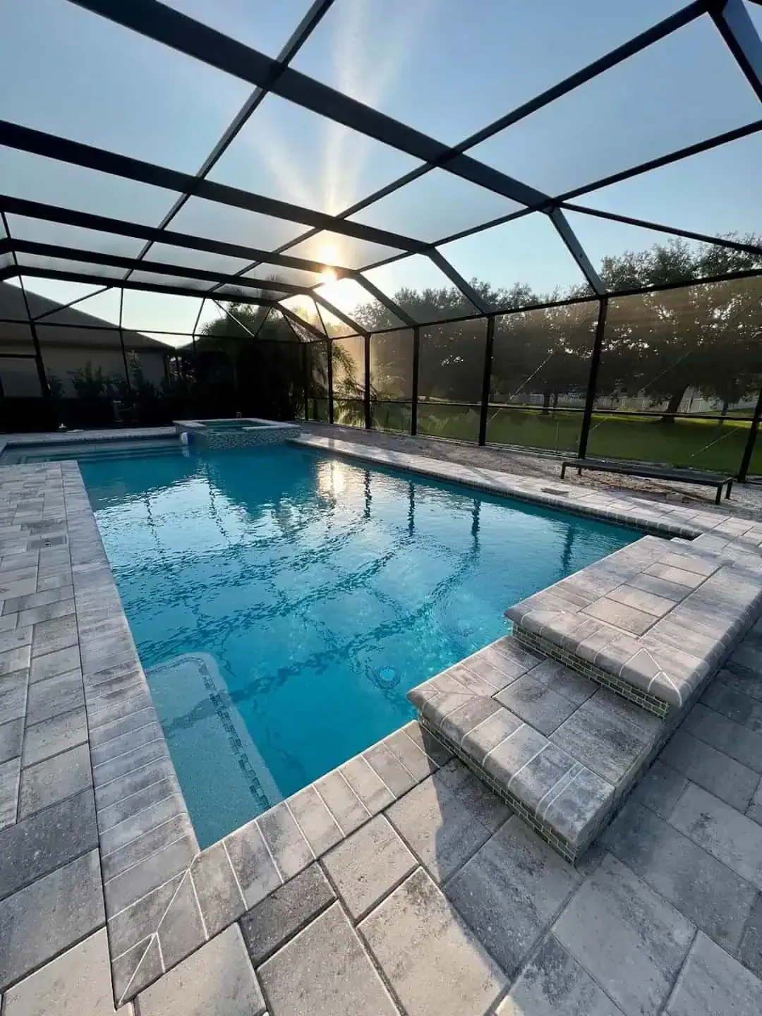 Screened backyard pool with stone deck and steps, sunlight reflecting on blue water