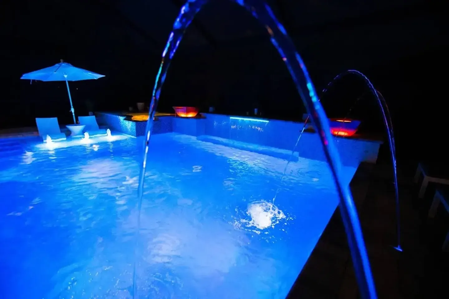 Blue-lit nighttime swimming pool with a few lounge chairs and a patio umbrella.