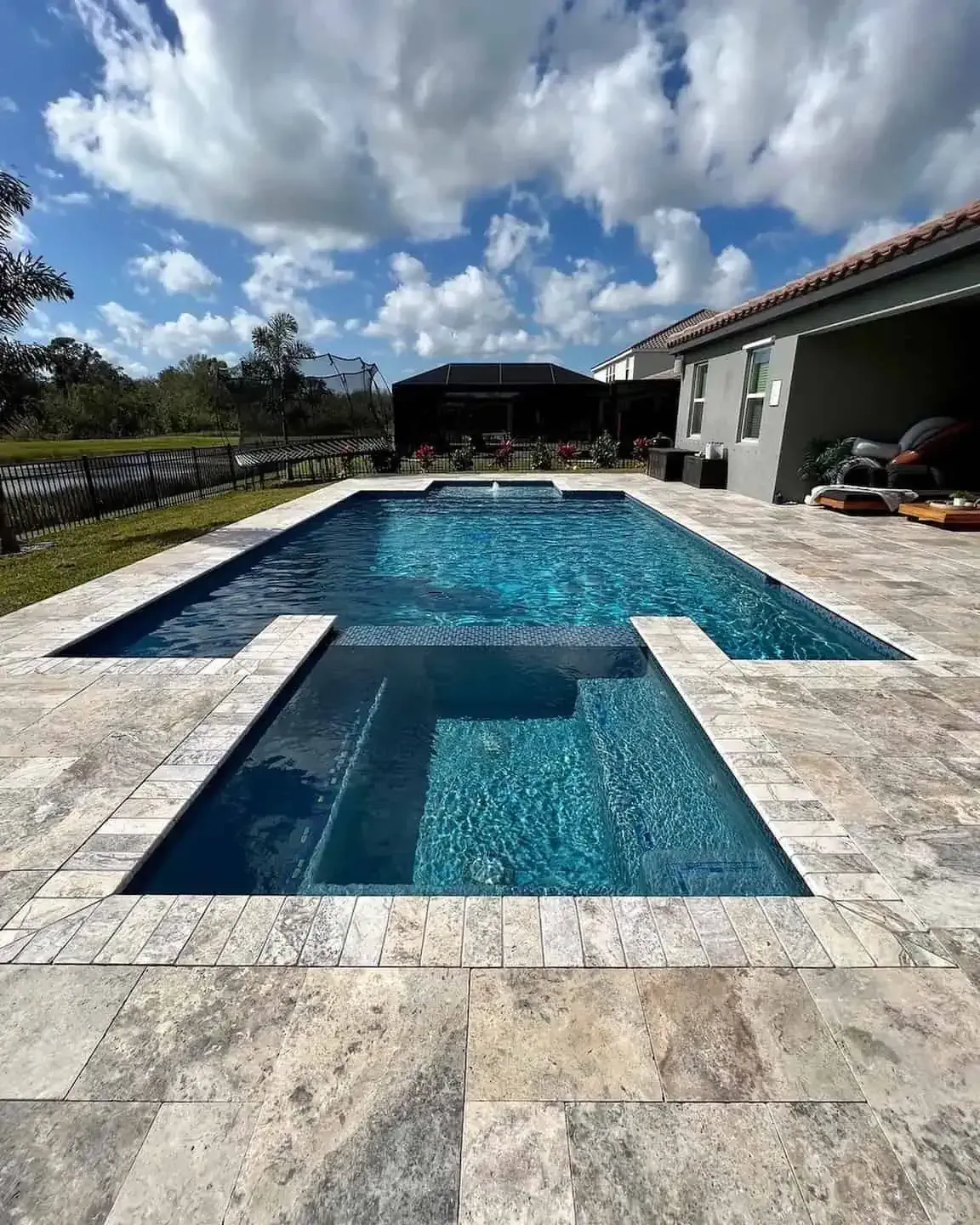 Modern backyard pool with blue water, stone deck, lounge chairs, and a partly cloudy sky