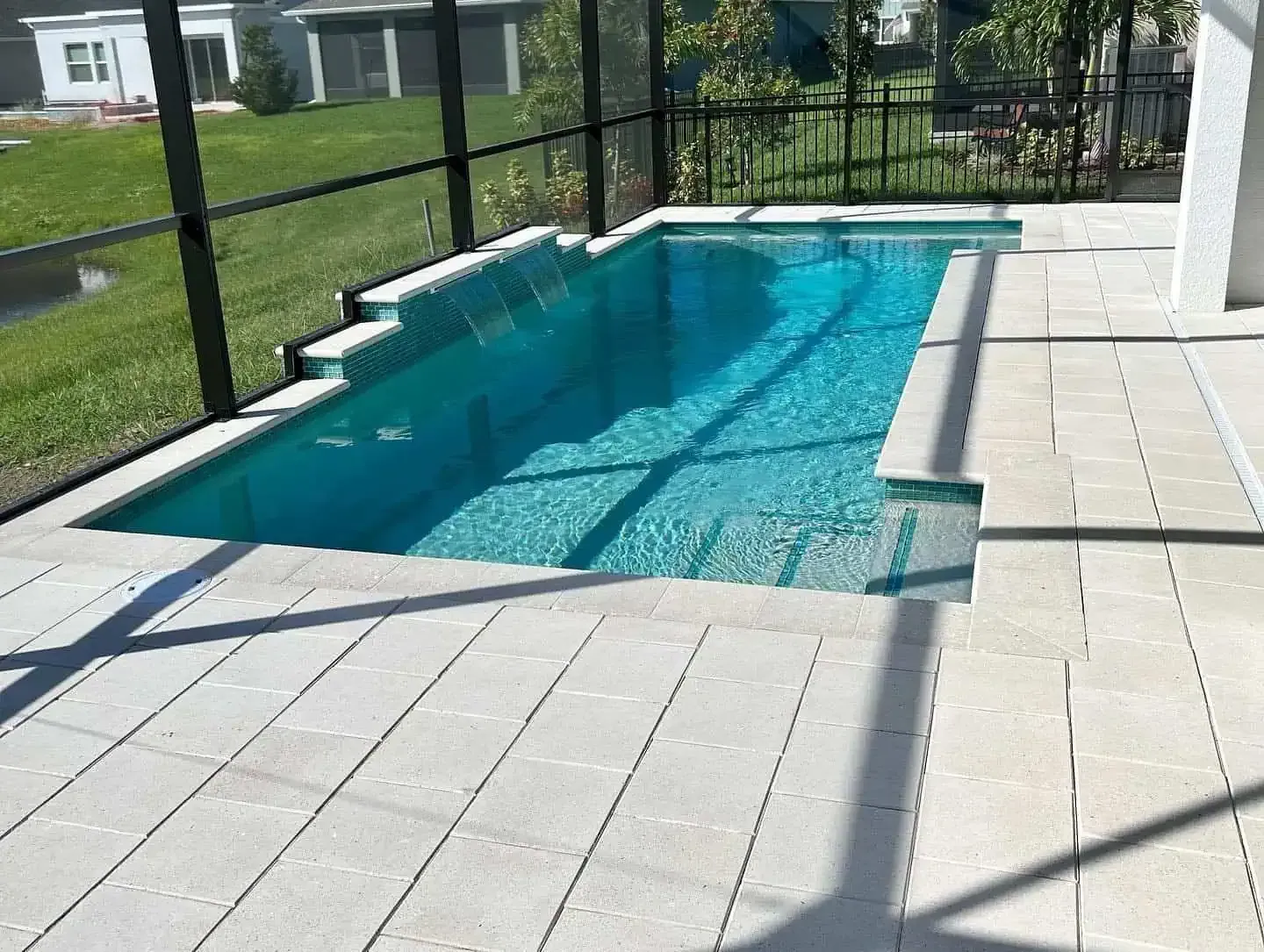 Rectangular turquoise pool beside a screened patio with light stone deck and grassy yard beyond