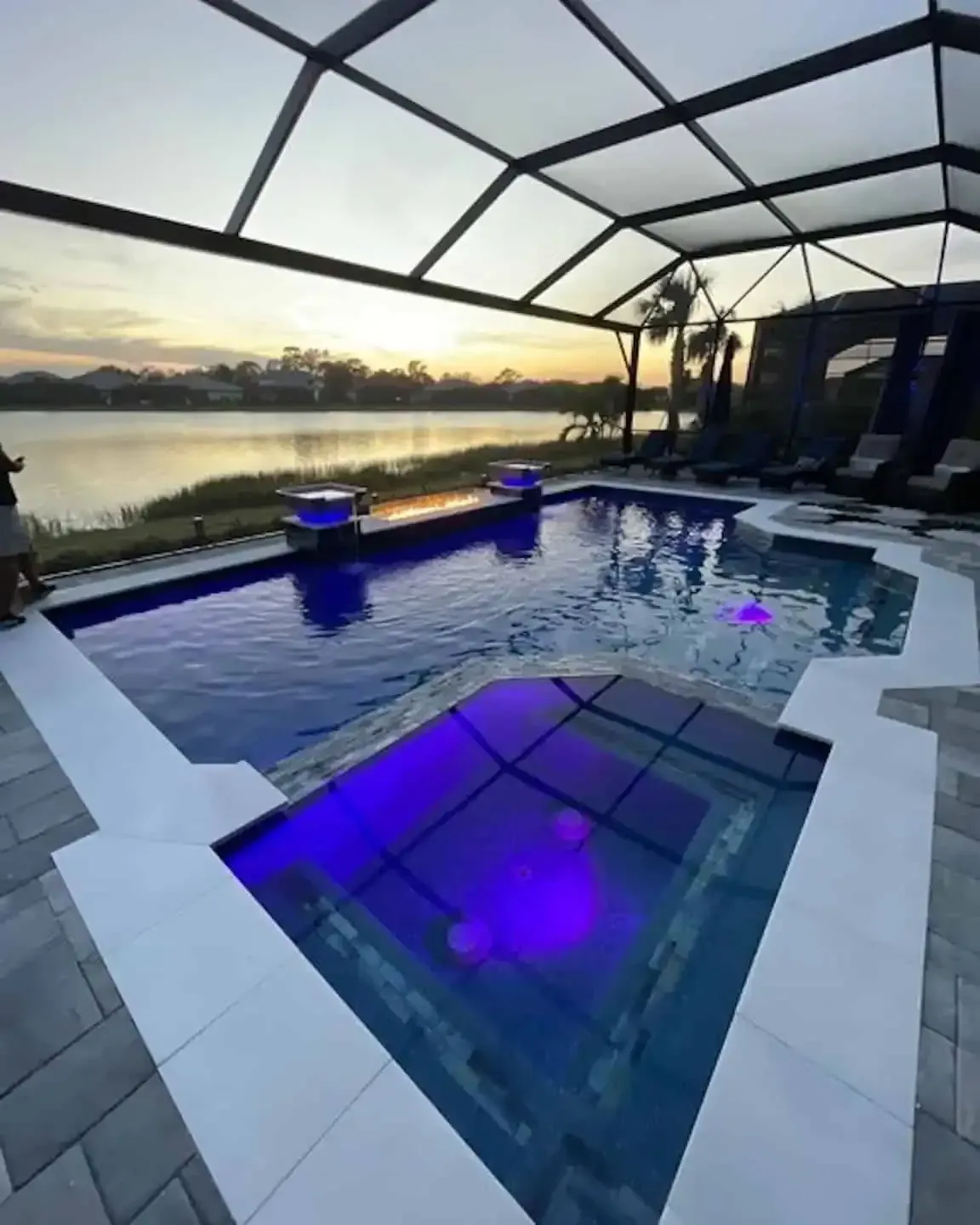 Covered backyard pool and hot tub at sunset with blue-purple lighting and a lake view