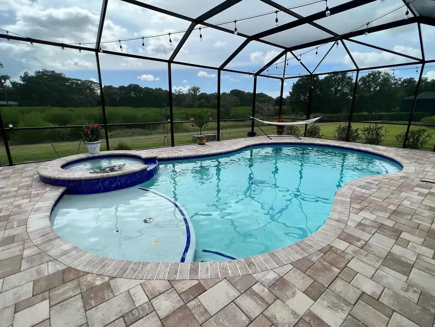 Screened backyard pool with a brick patio, hot tub, and grassy landscape beyond.