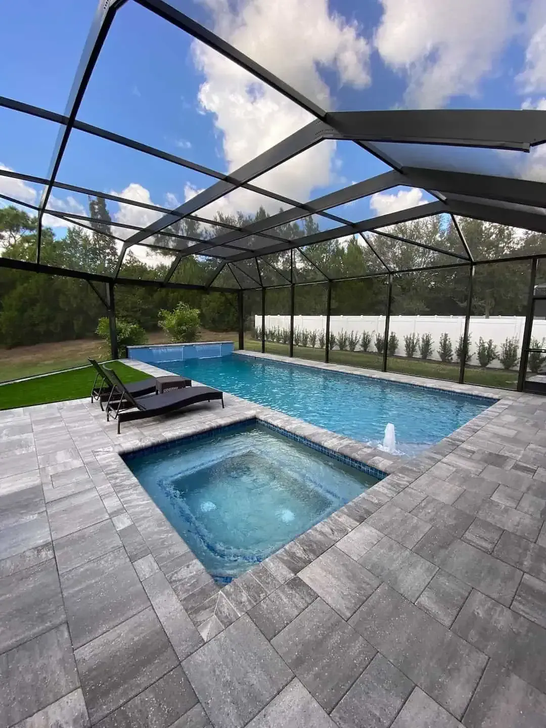 Screened backyard pool and spa with gray pavers, lounge chairs, and blue sky overhead