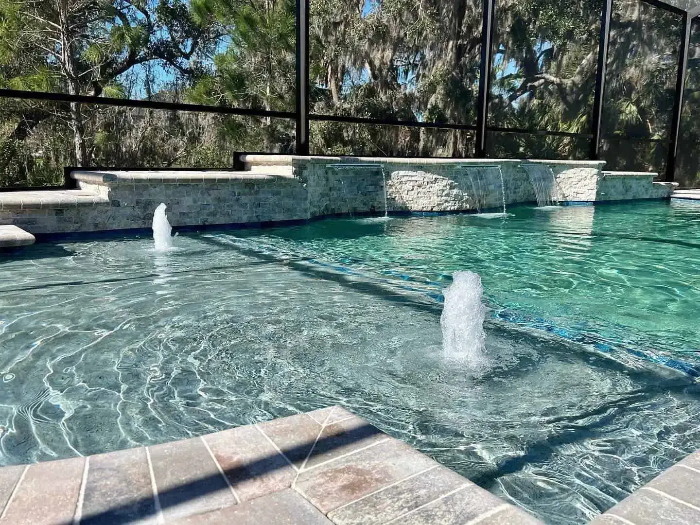 Sunlit outdoor pool with two bubbling fountains and a stone deck by a screened enclosure