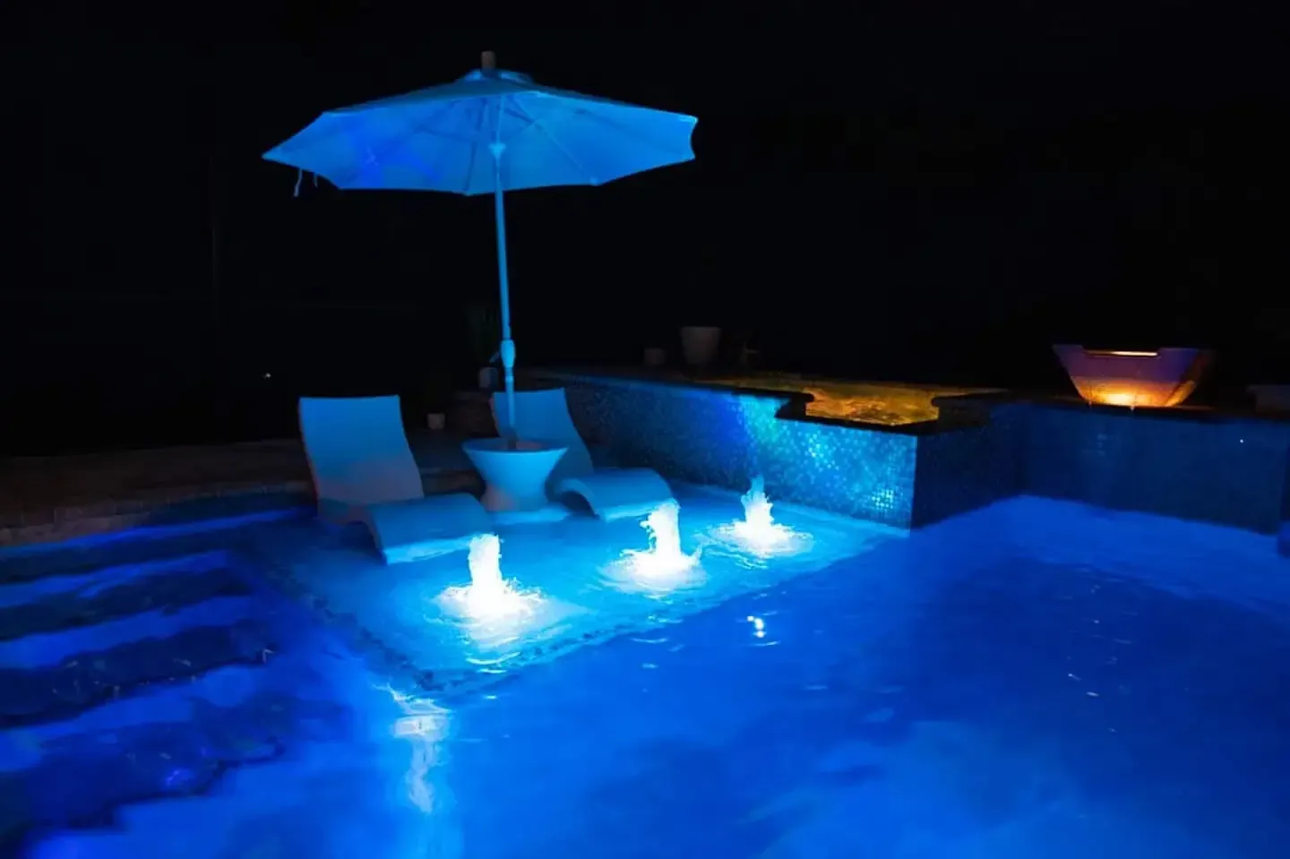 Blue-lit swimming pool at night with a lounge chair, umbrella, and glowing water jets
