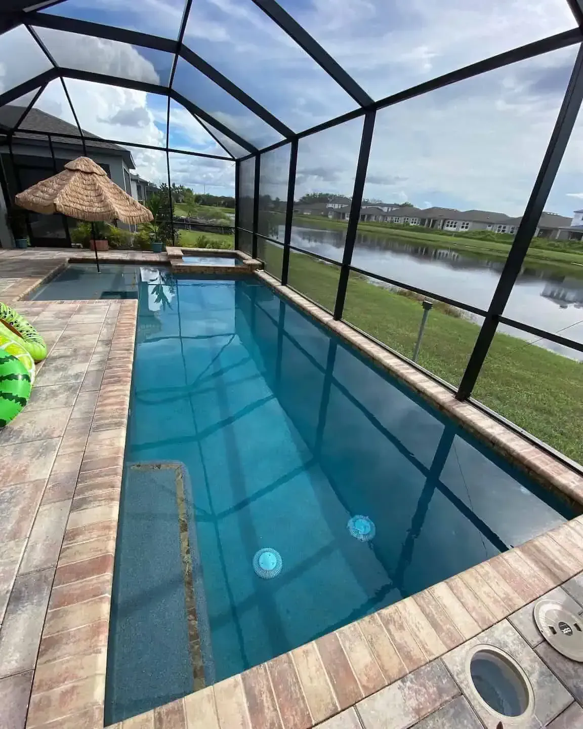 Screened backyard pool with blue water beside a canal, paver deck, and thatched gazebo under a cloudy sky