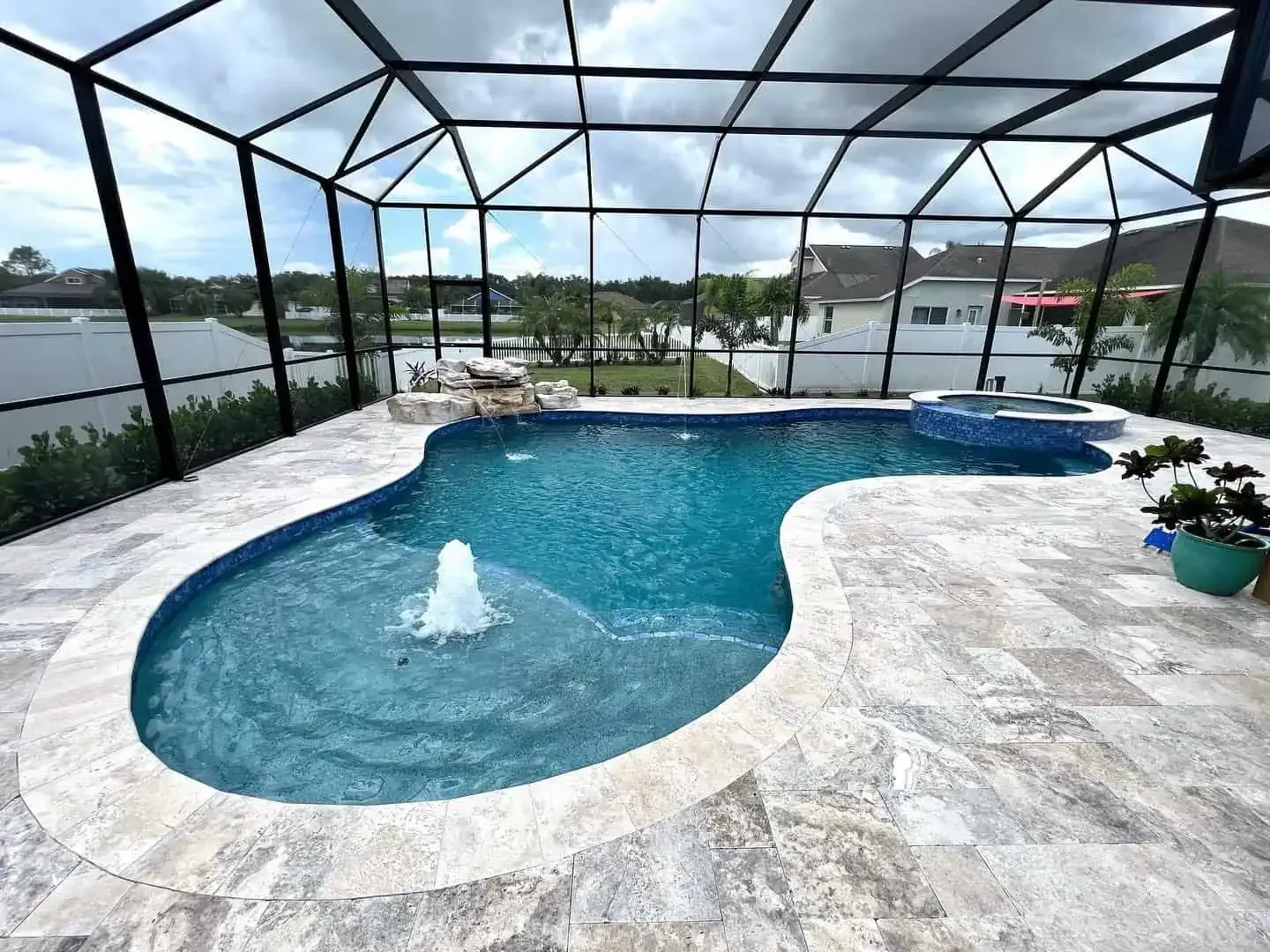 Screened-in backyard pool with spa and fountain, surrounded by light stone decking under a cloudy sky