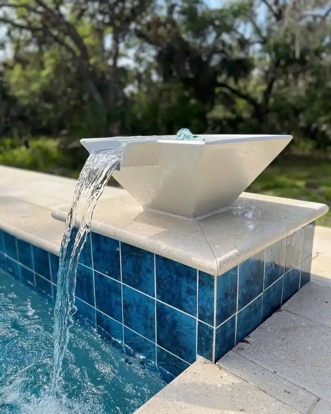 White fountain pouring water into a blue-tiled pool beside a beige stone ledge