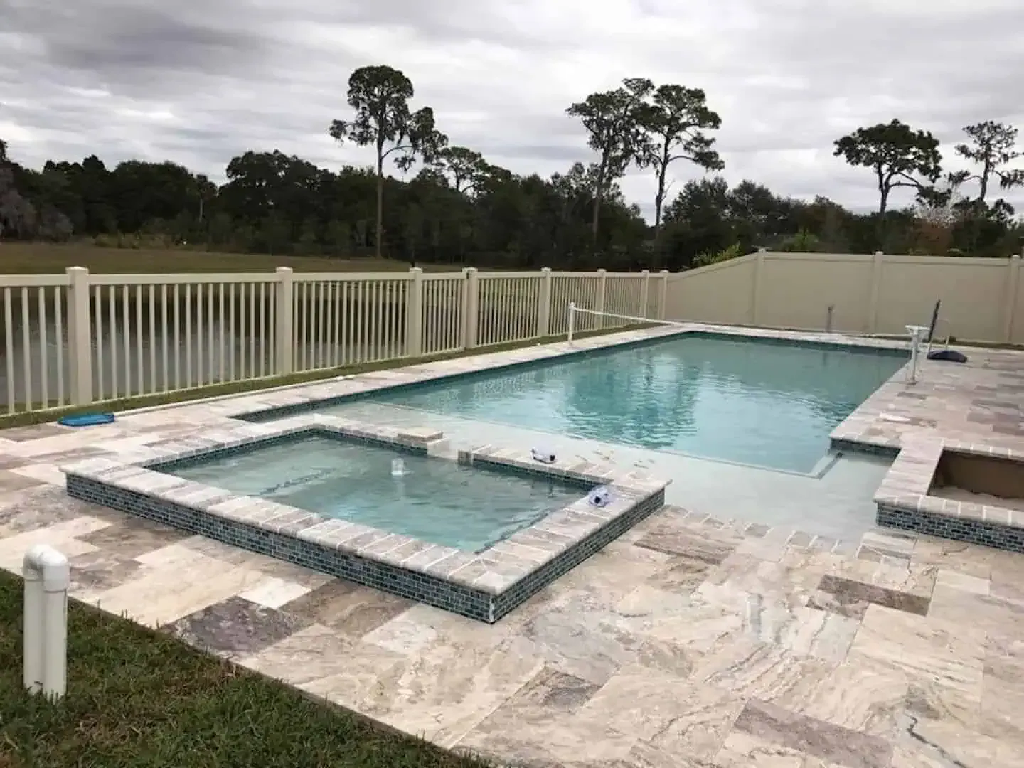 Backyard pool with a raised spa, stone patio, and white fence beside trees and lawn