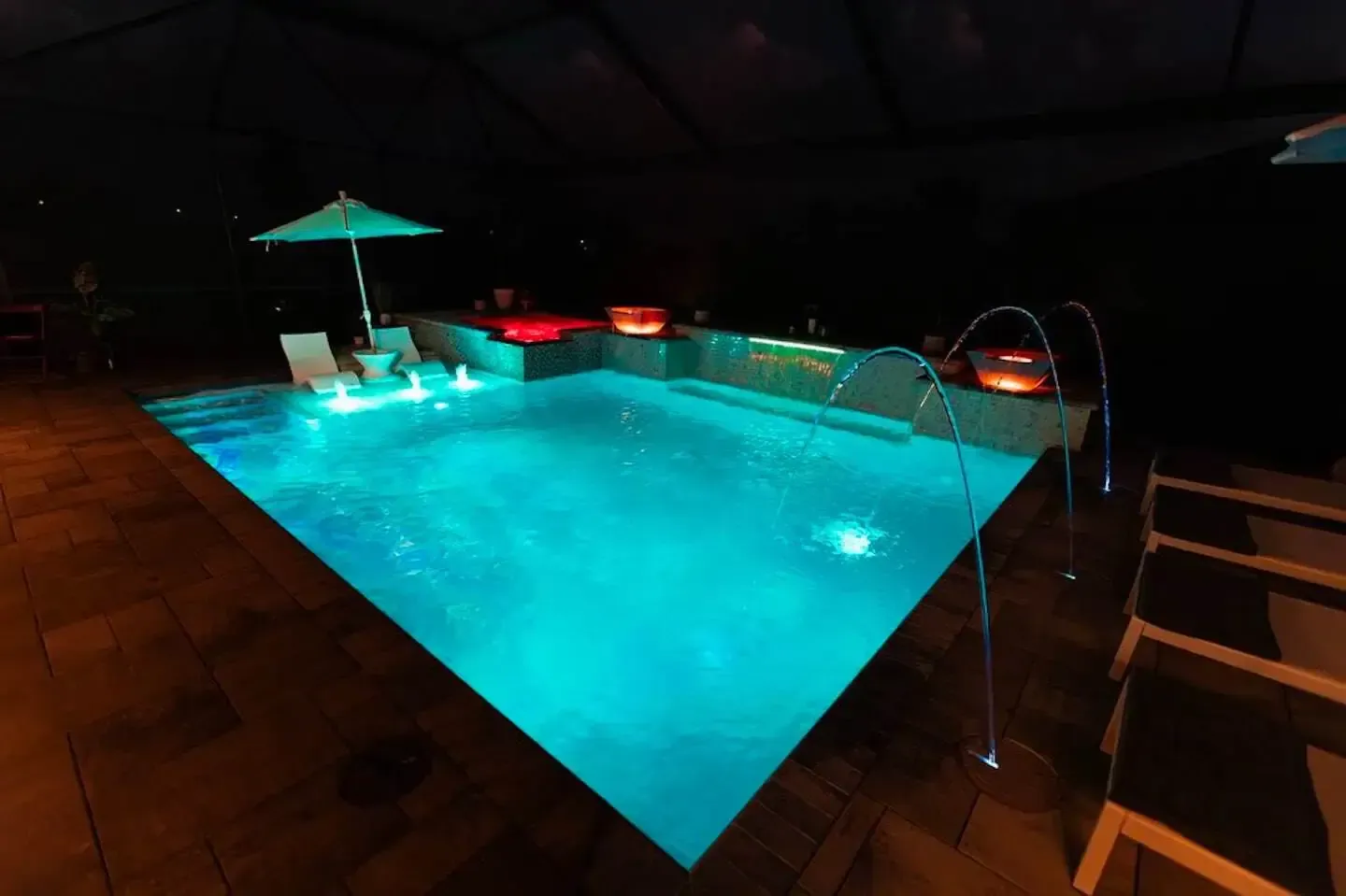 Illuminated indoor swimming pool with turquoise water, umbrella, lounge chairs, and stairs in a dark setting