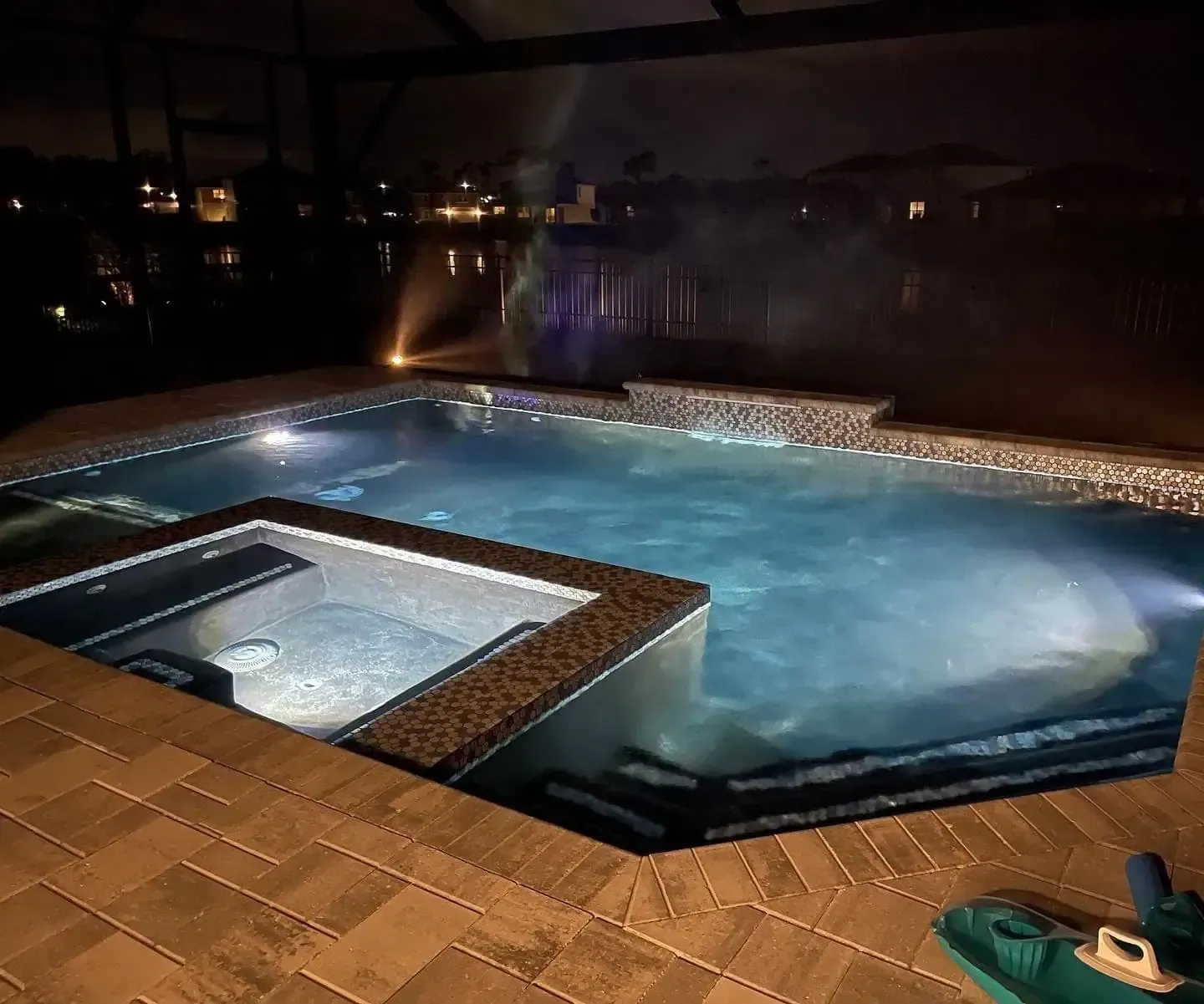 Nighttime backyard pool and hot tub with illuminated water and stone deck.