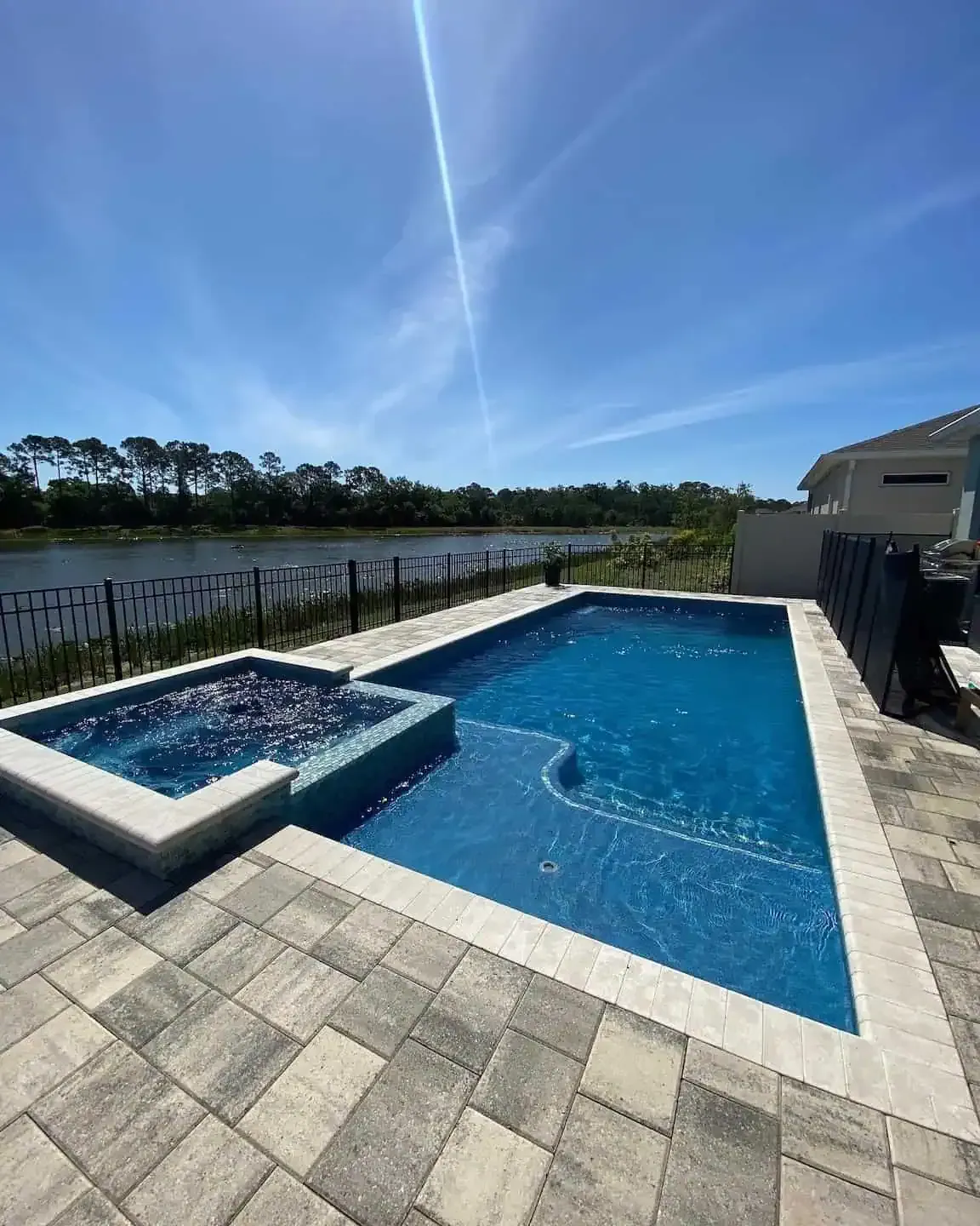 Backyard pool and hot tub with blue water, stone patio, and lake view under a clear sky