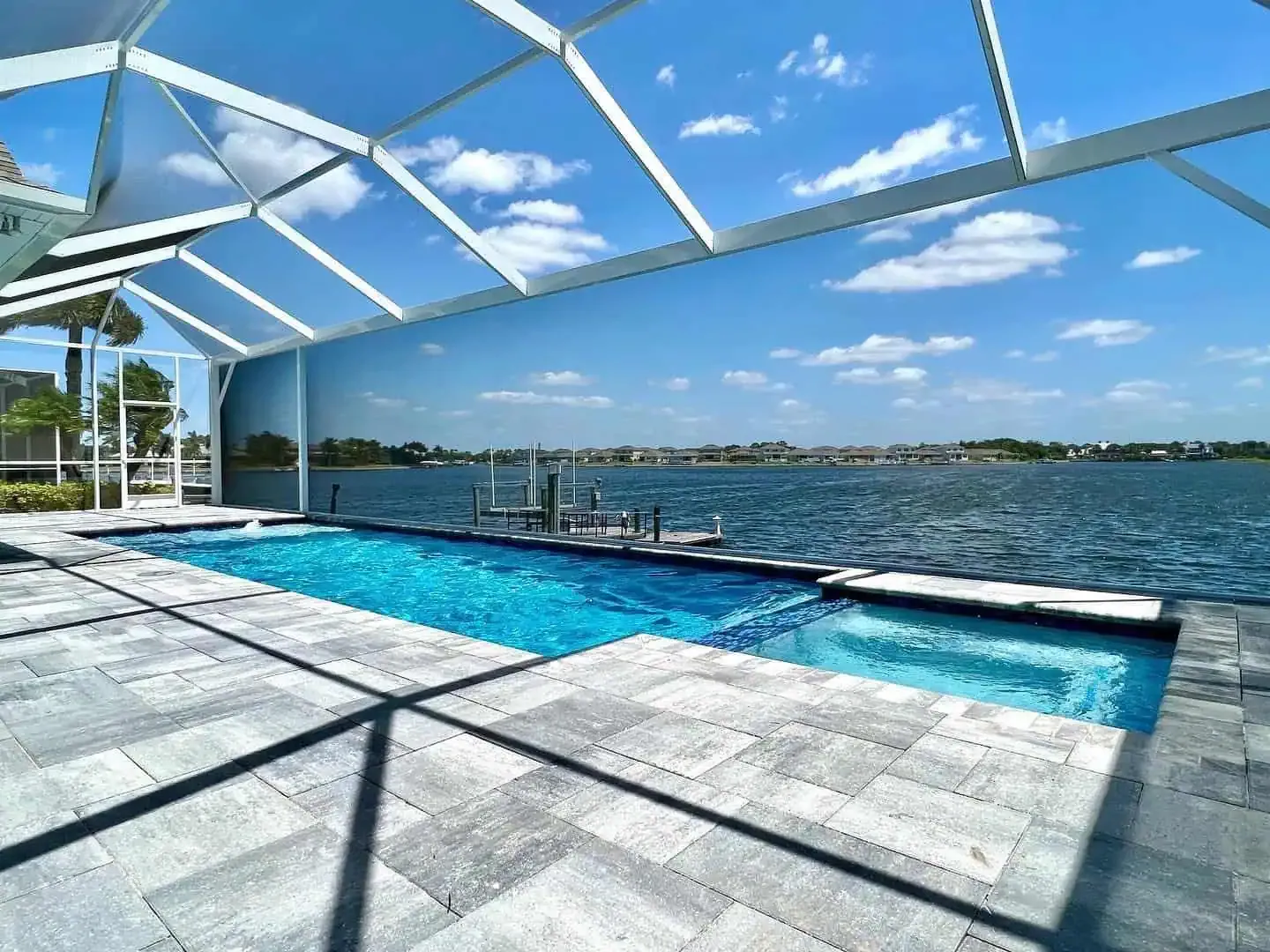 Screened outdoor pool beside a waterfront, with blue water, white deck, and clear sky above.