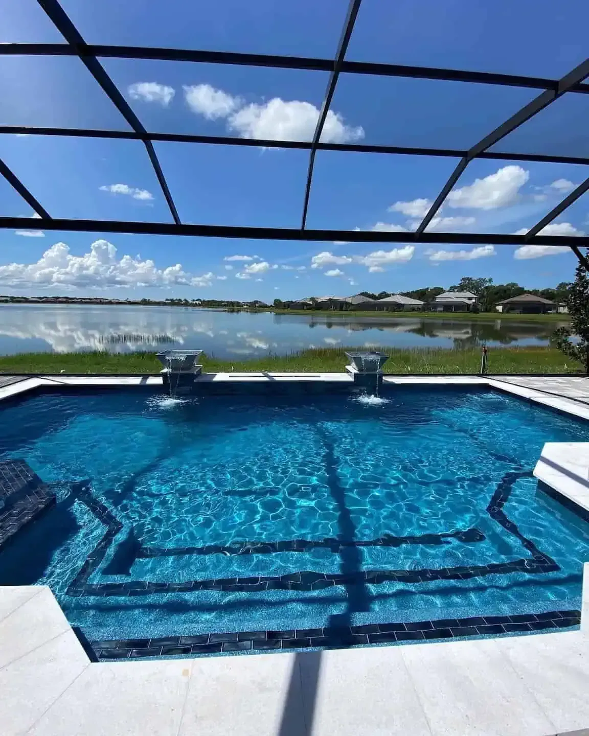 Blue infinity pool overlooking a lake under a screened patio, with lounge chairs and a cloudy sky