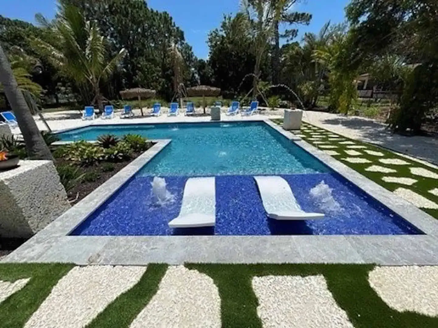 Modern backyard pool with sun loungers, blue water, and striped stone walkway amid lush greenery