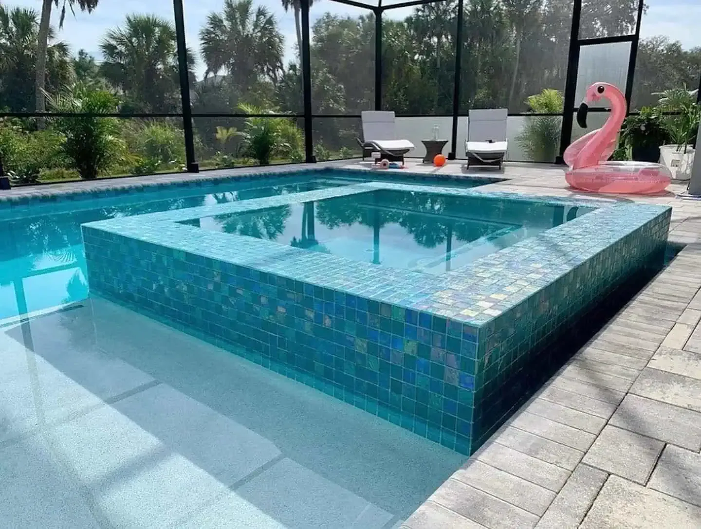 Modern backyard pool with raised tiled spa, lounge chairs, and a pink flamingo float by a screened enclosure