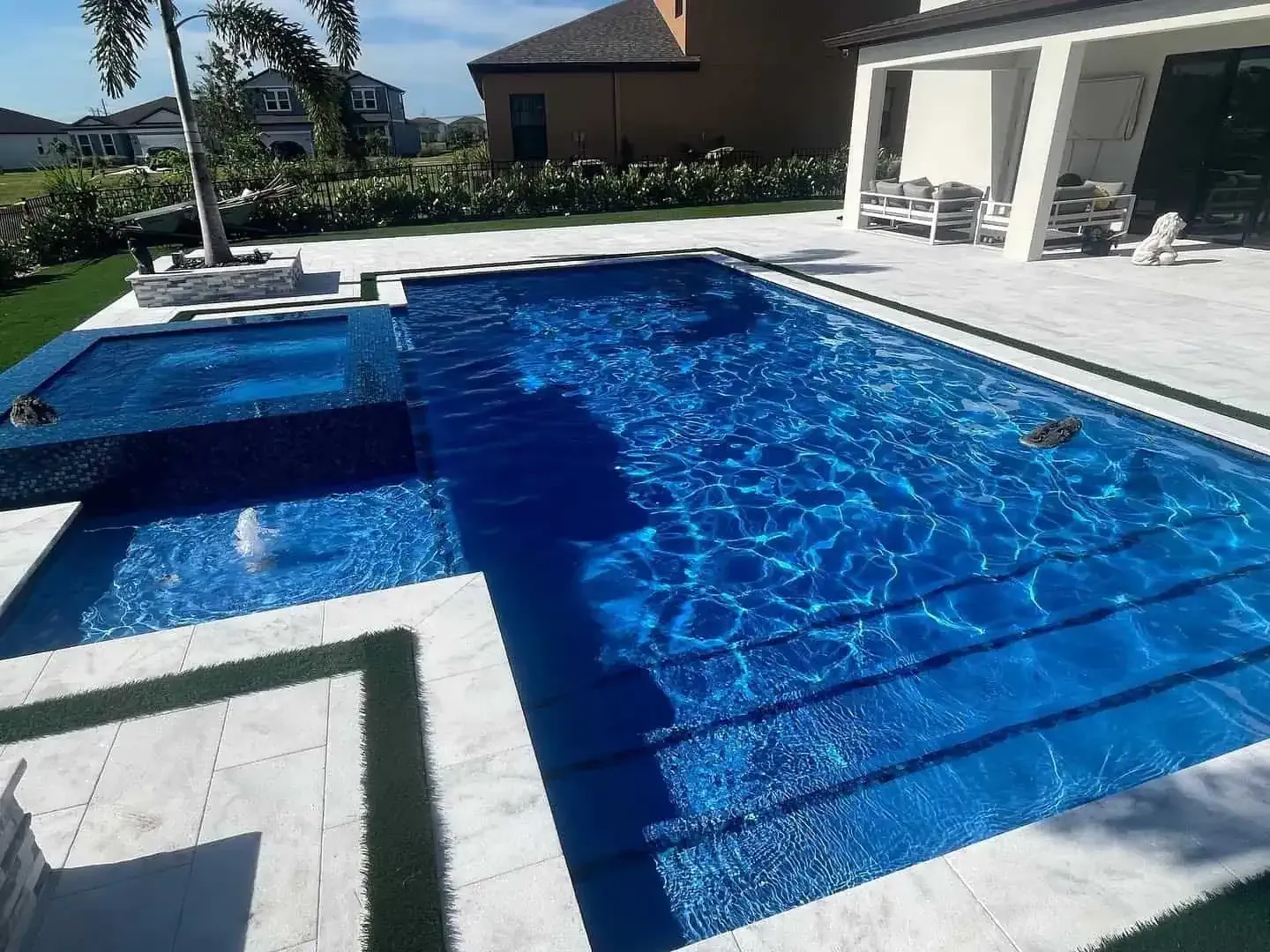 Blue tiled backyard swimming pool with a shallow ledge and white patio beside a house