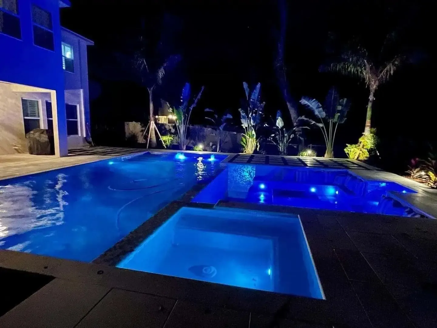 Blue-lit backyard pool and hot tub at night beside a house, with palm trees and landscaping