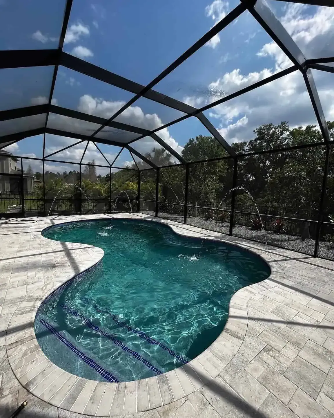 Screened-in backyard pool with a curved turquoise pool and light stone deck under a blue sky