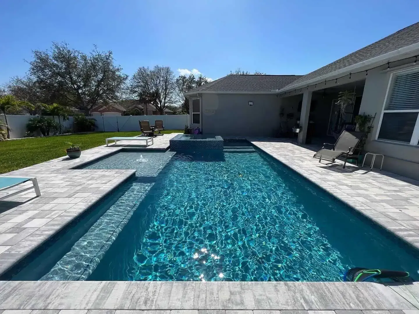 Backyard swimming pool with a raised spa, stone deck, and patio beside a house on a sunny day