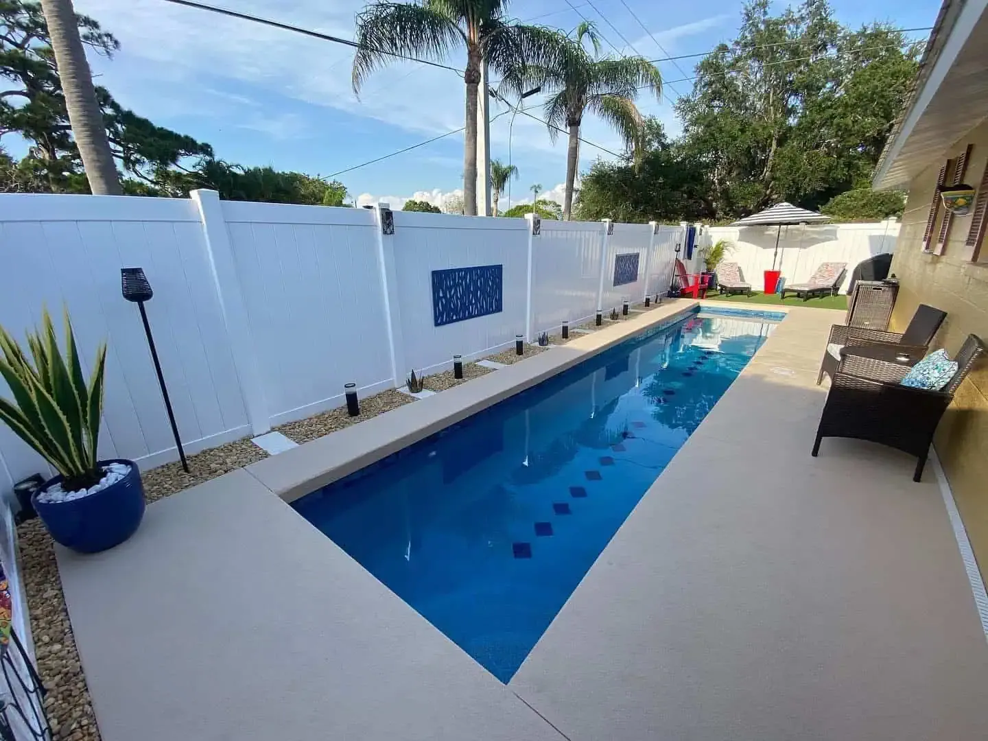Narrow backyard pool with blue water, white walls, lounge chairs, and potted plants on a sunny day