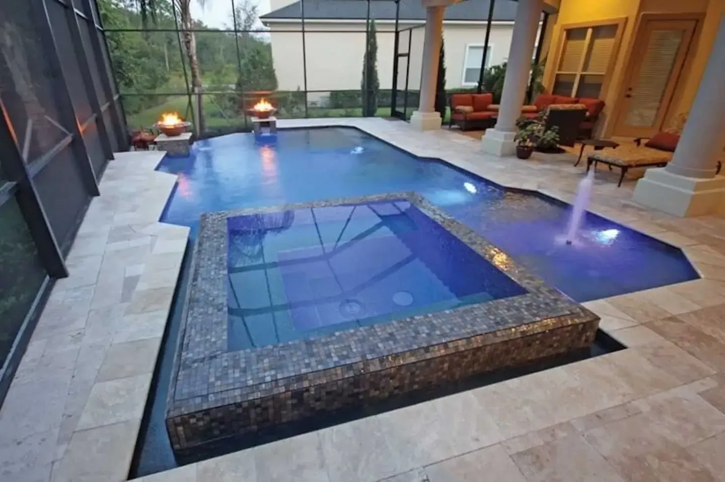 Blue-lit backyard pool with a raised spa and water jets on a stone patio at dusk