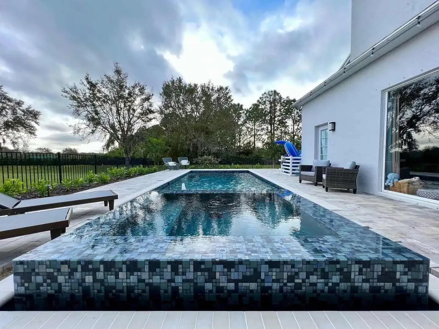 Modern backyard infinity pool with mosaic tile, white patio, lounge chairs, and cloudy sky