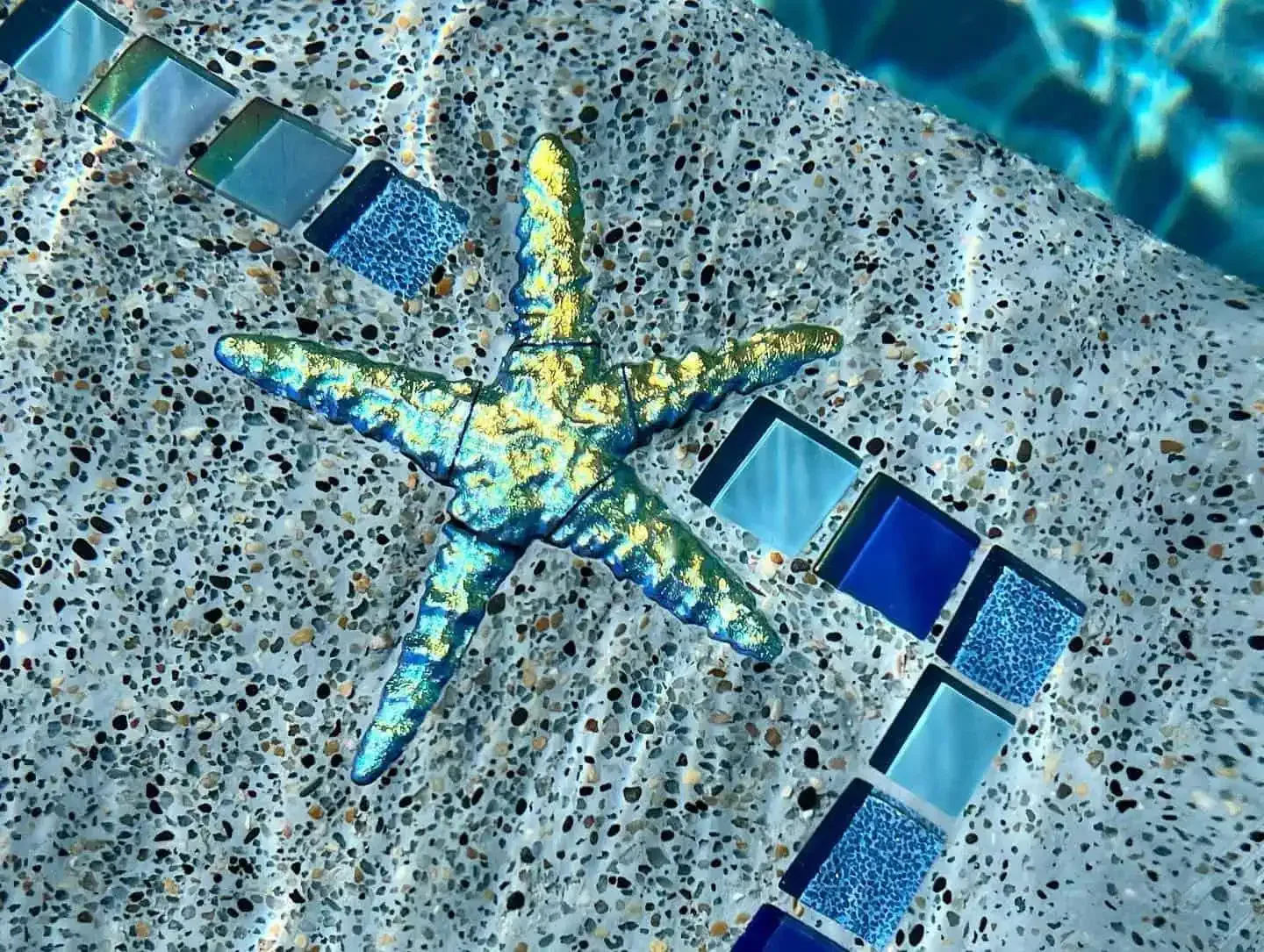 A blue-green starfish mosaic in a speckled tiled pool floor with a curved line of blue tiles nearby
