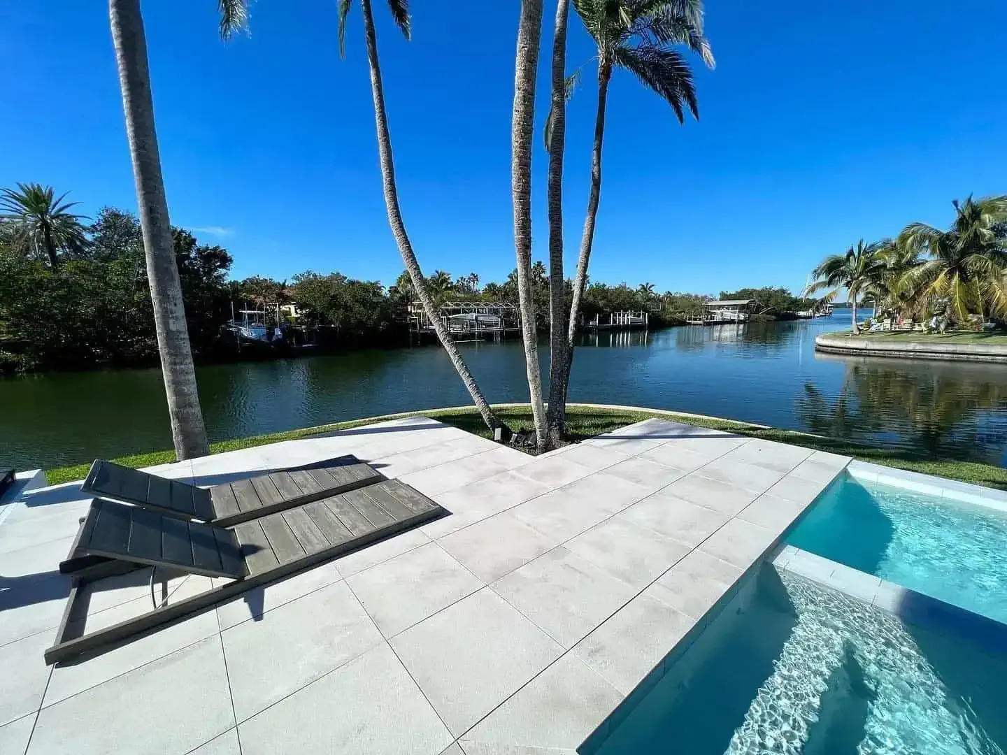 Sunlit backyard pool deck with lounge chairs, palm trees, and a canal view