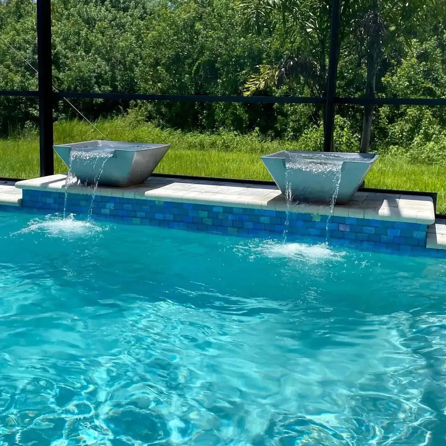 Blue swimming pool with two metal water features on the deck and a green lawn in the background
