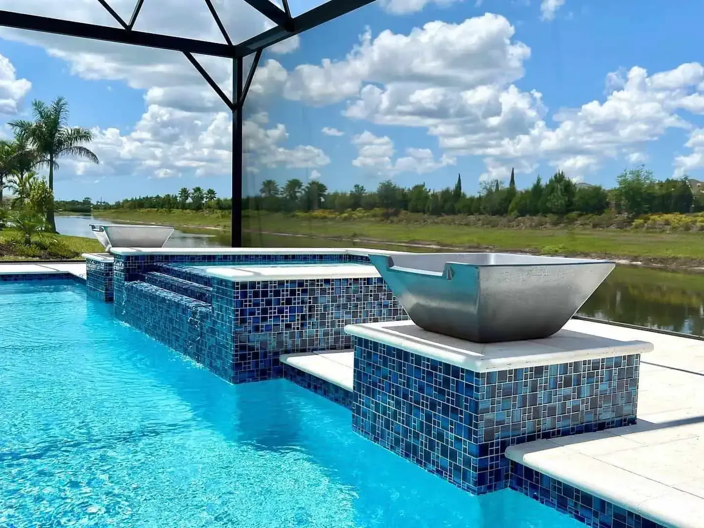 Infinity pool with blue mosaic tiles, white lounge table, and a canopy overlooking a grassy lakeside view.