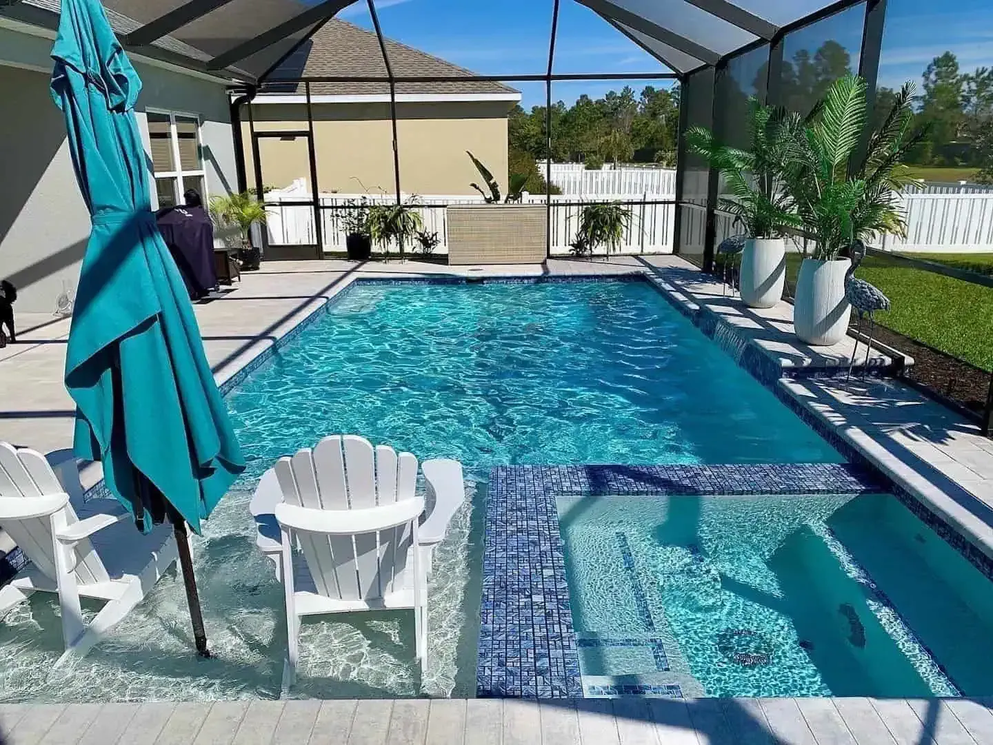 Backyard pool with screened enclosure, hot tub, white chairs, and teal umbrella on a sunny day