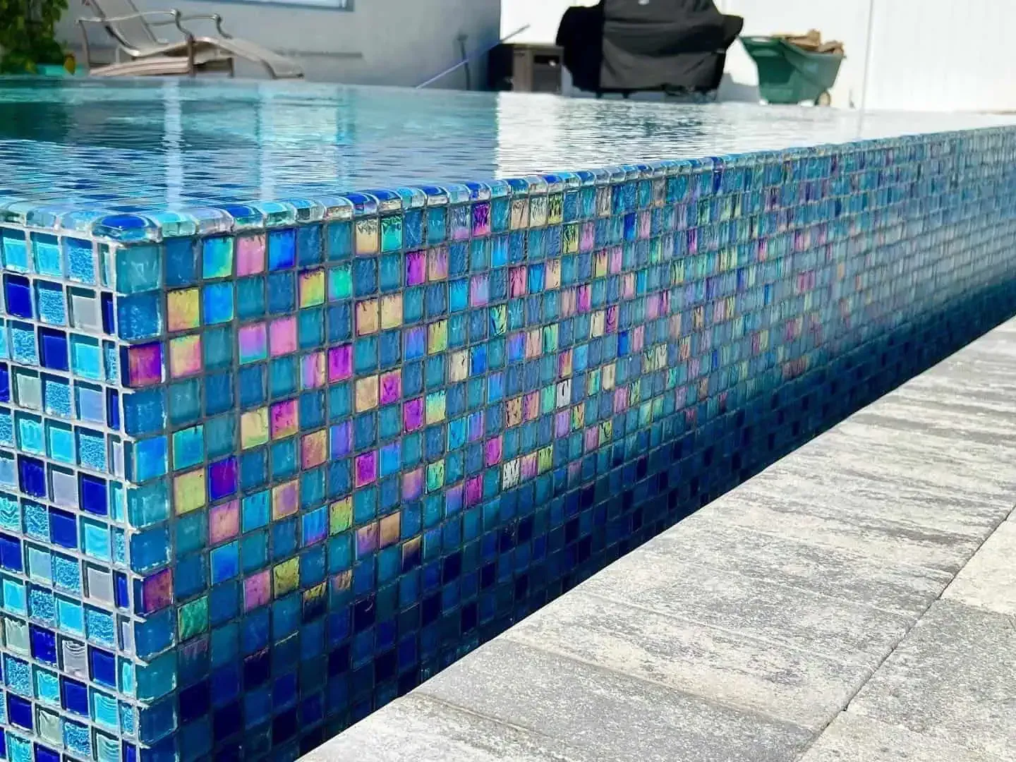 Colorful mosaic-tiled swimming pool edge beside clear blue water and gray stone deck
