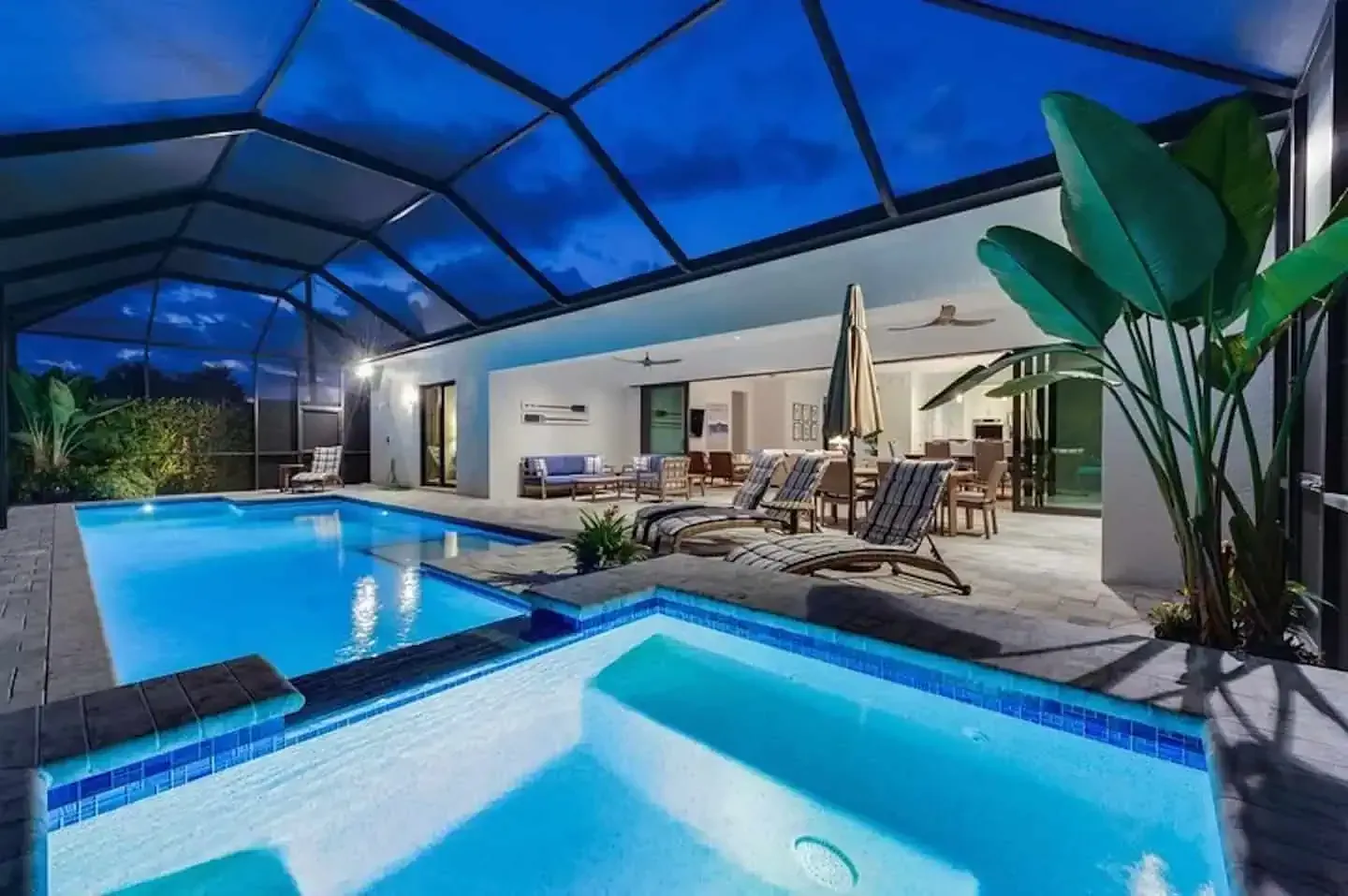 Modern poolside patio at night with blue-lit pool, spa, lounge chairs, and covered seating area