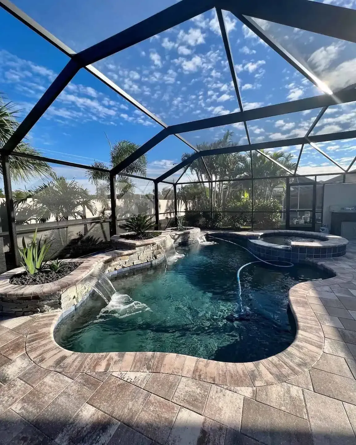 Screened backyard pool with stone deck, spa, and clear blue water under a bright sky