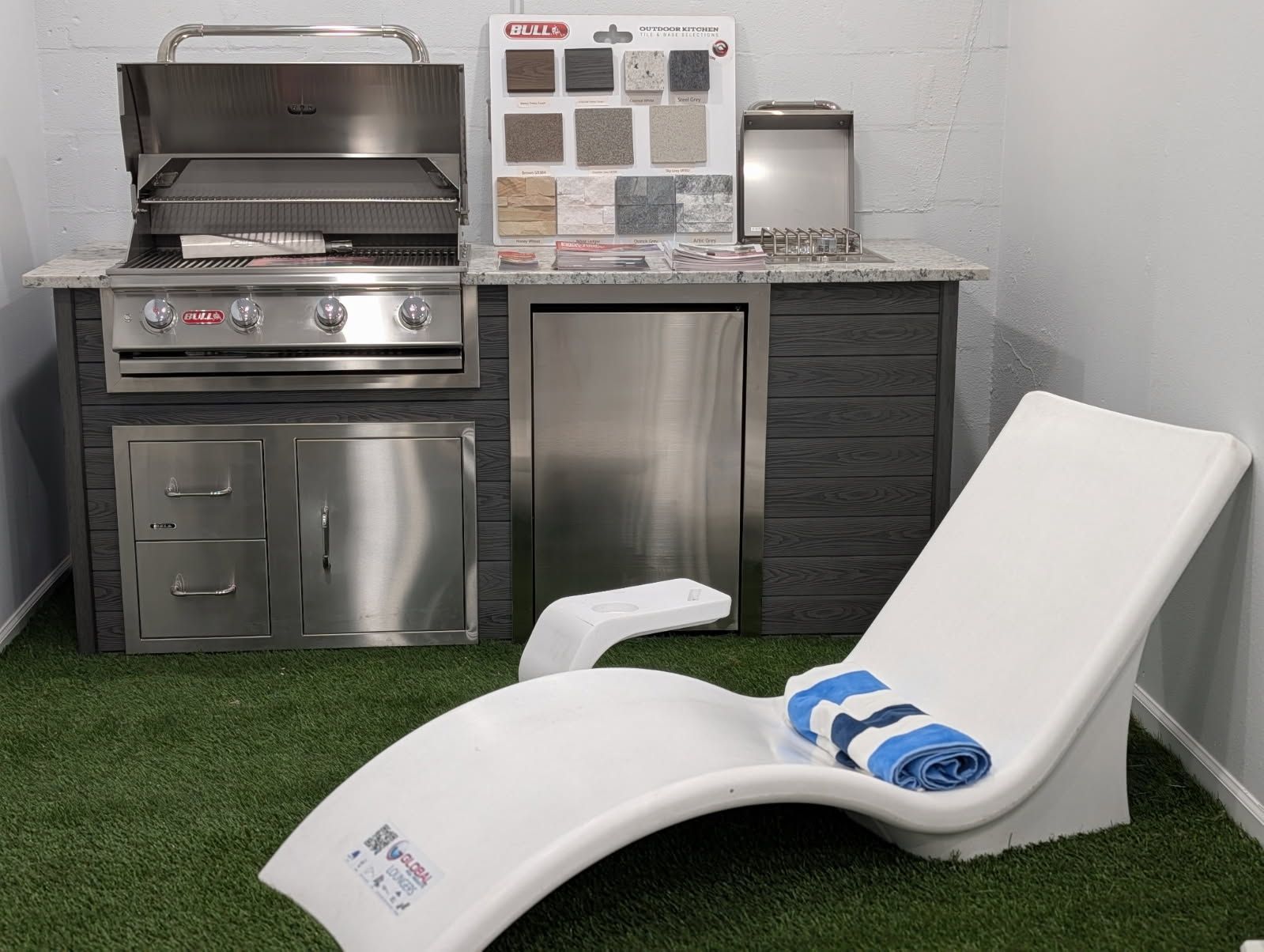 Patio grill setup with stainless steel BBQ, outdoor kitchen cabinets, and two white chaise lounges with blue towels.