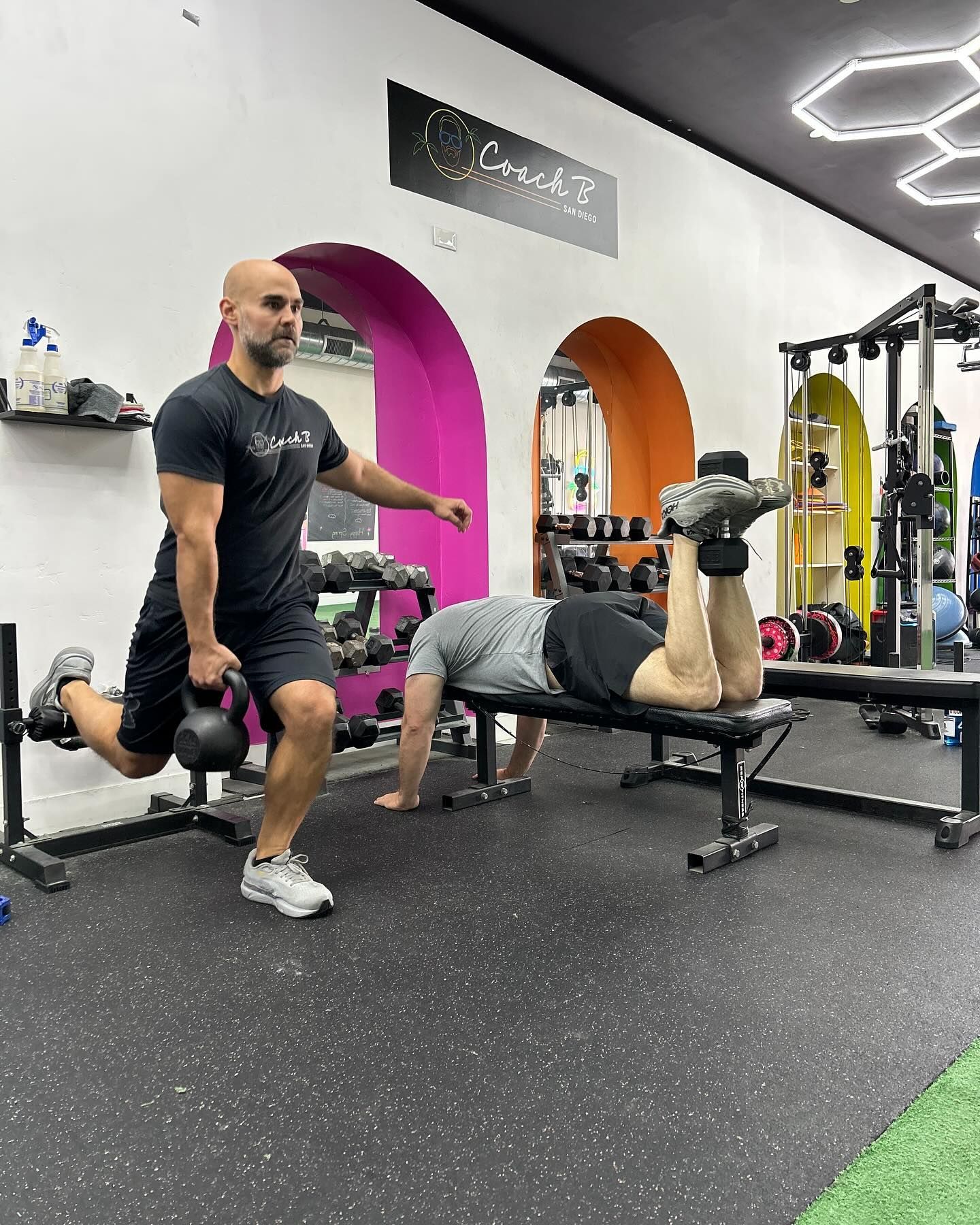 A man is standing next to a man laying on a bench in a gym.