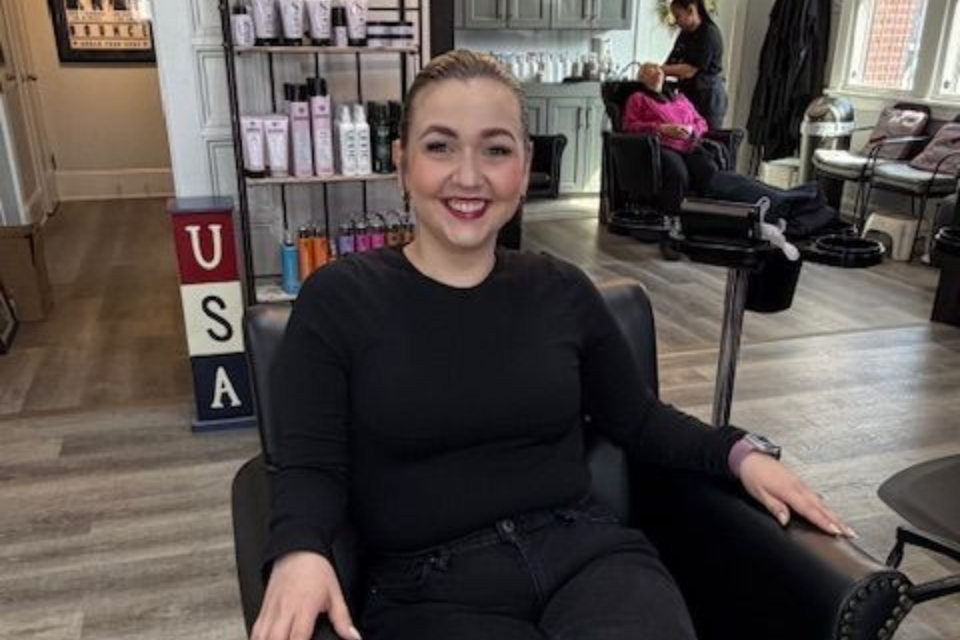 Smiling person seated in a salon chair, with mirrors, hair products, and styling chairs in the background.