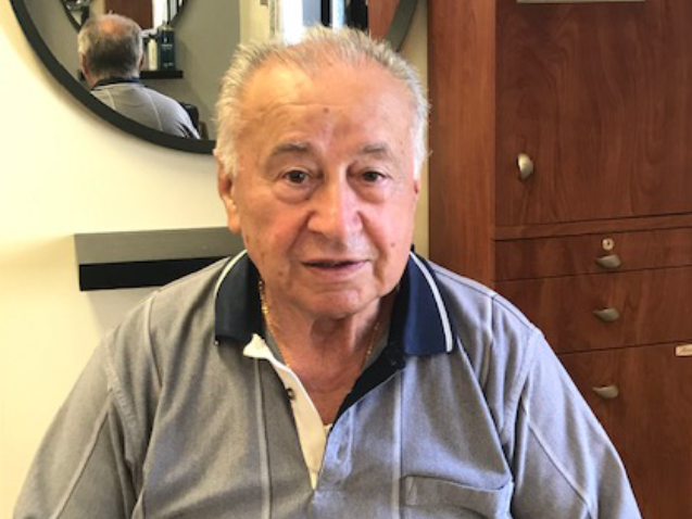 Older man in a gray polo shirt sitting indoors, looking at the camera, with wood cabinets behind him