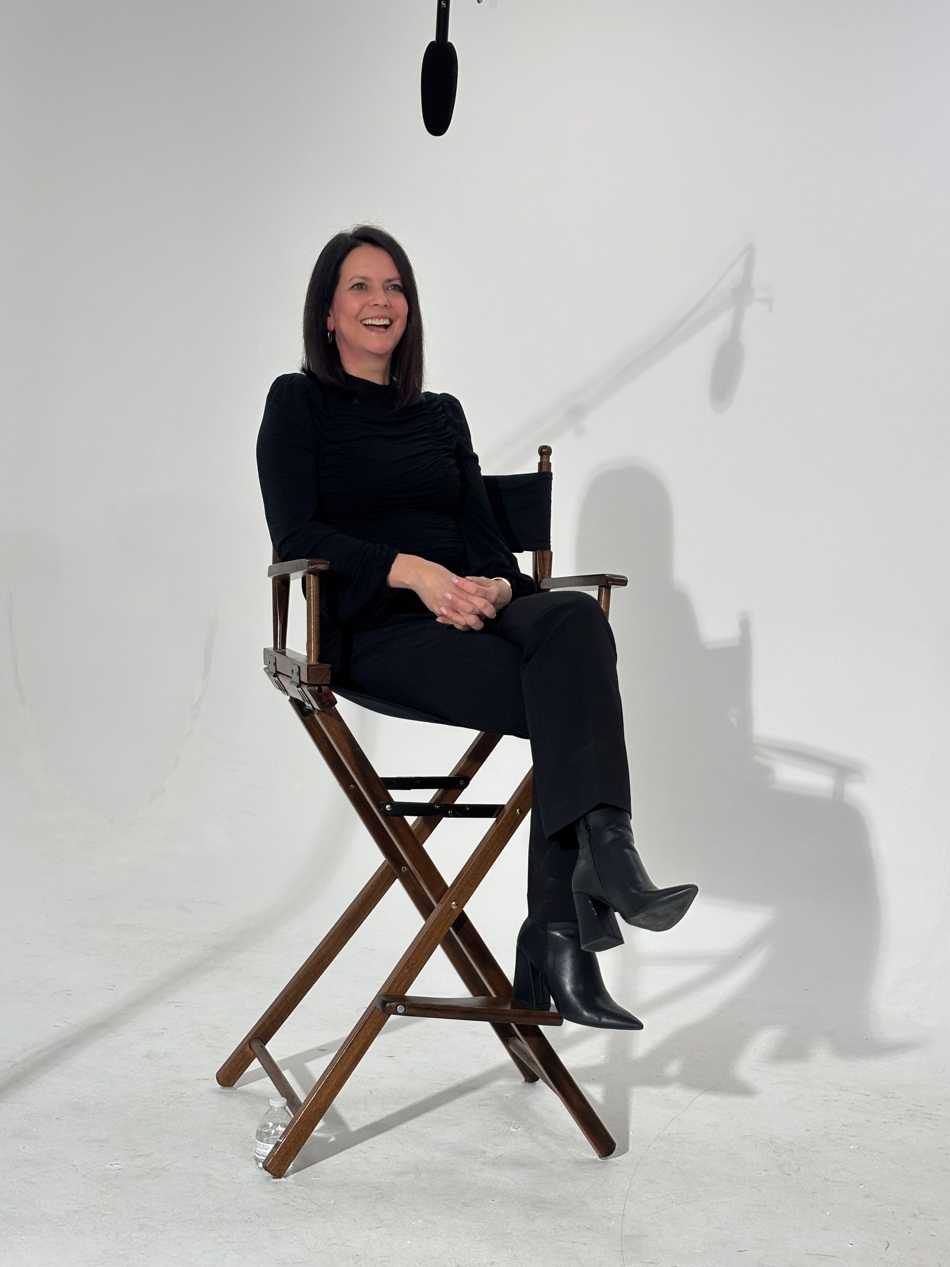 Woman seated on a wooden director’s chair in a white studio, wearing black clothes and boots.