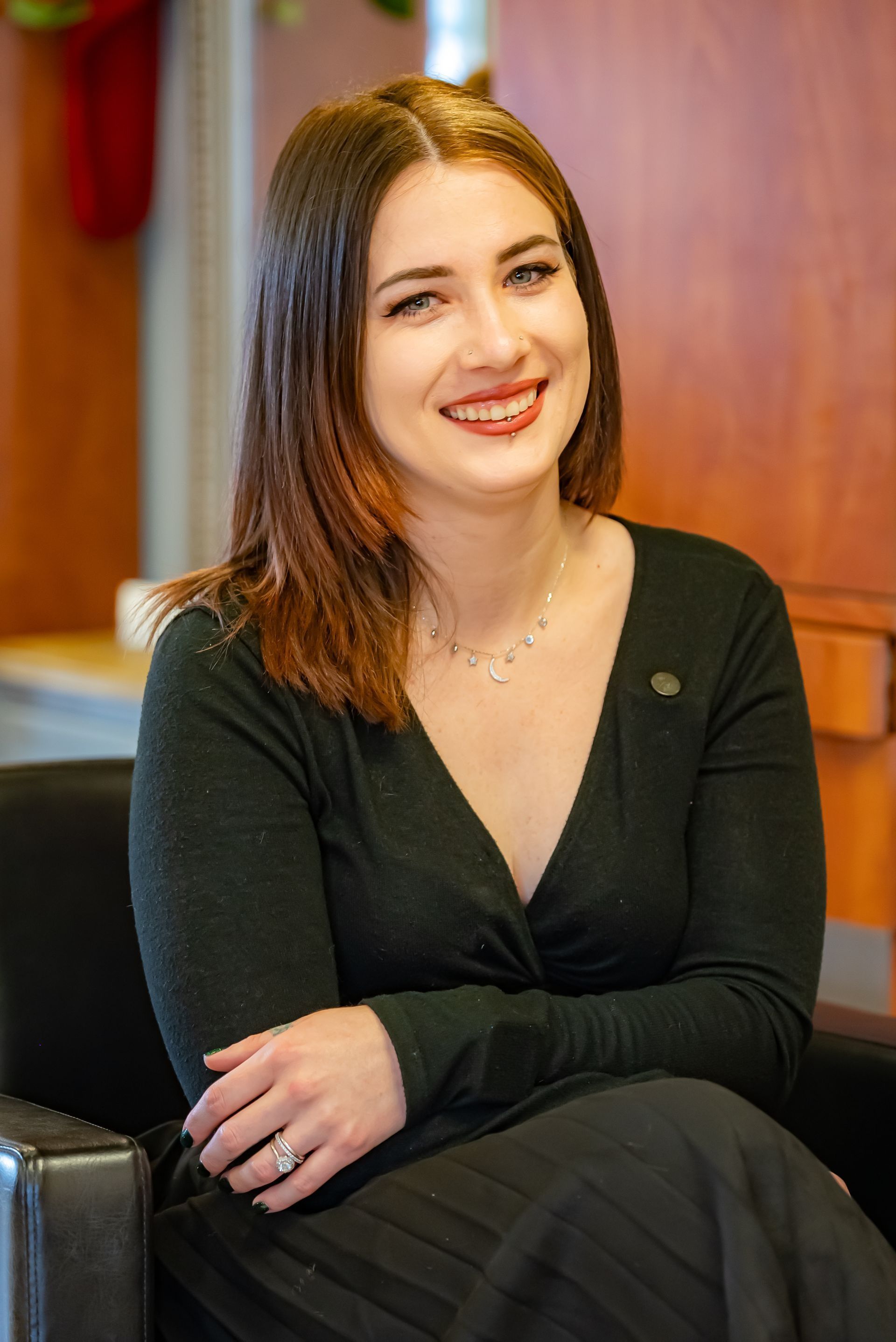 Smiling woman in a black dress seated indoors against a warm wood-paneled background