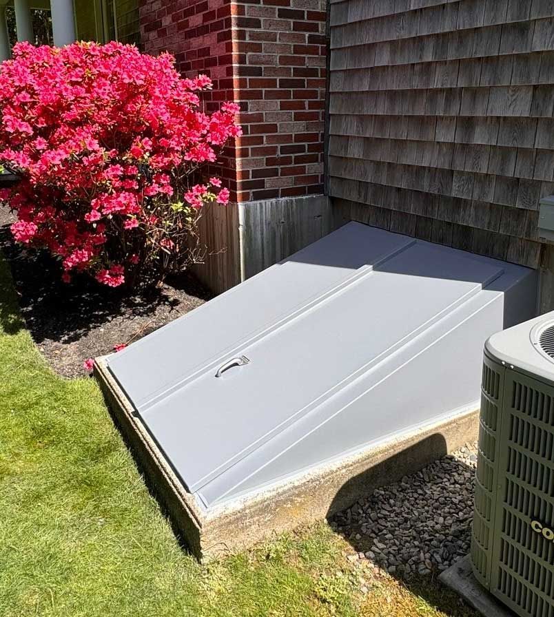 Gray cellar doors, set in concrete, next to a red-flowering bush and a brick house.