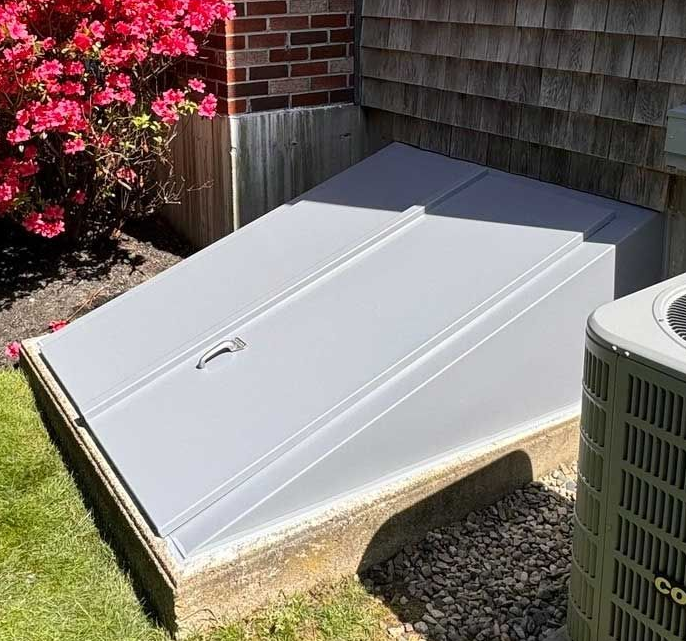 Gray cellar doors, set in concrete, next to a red-flowering bush and a brick house.