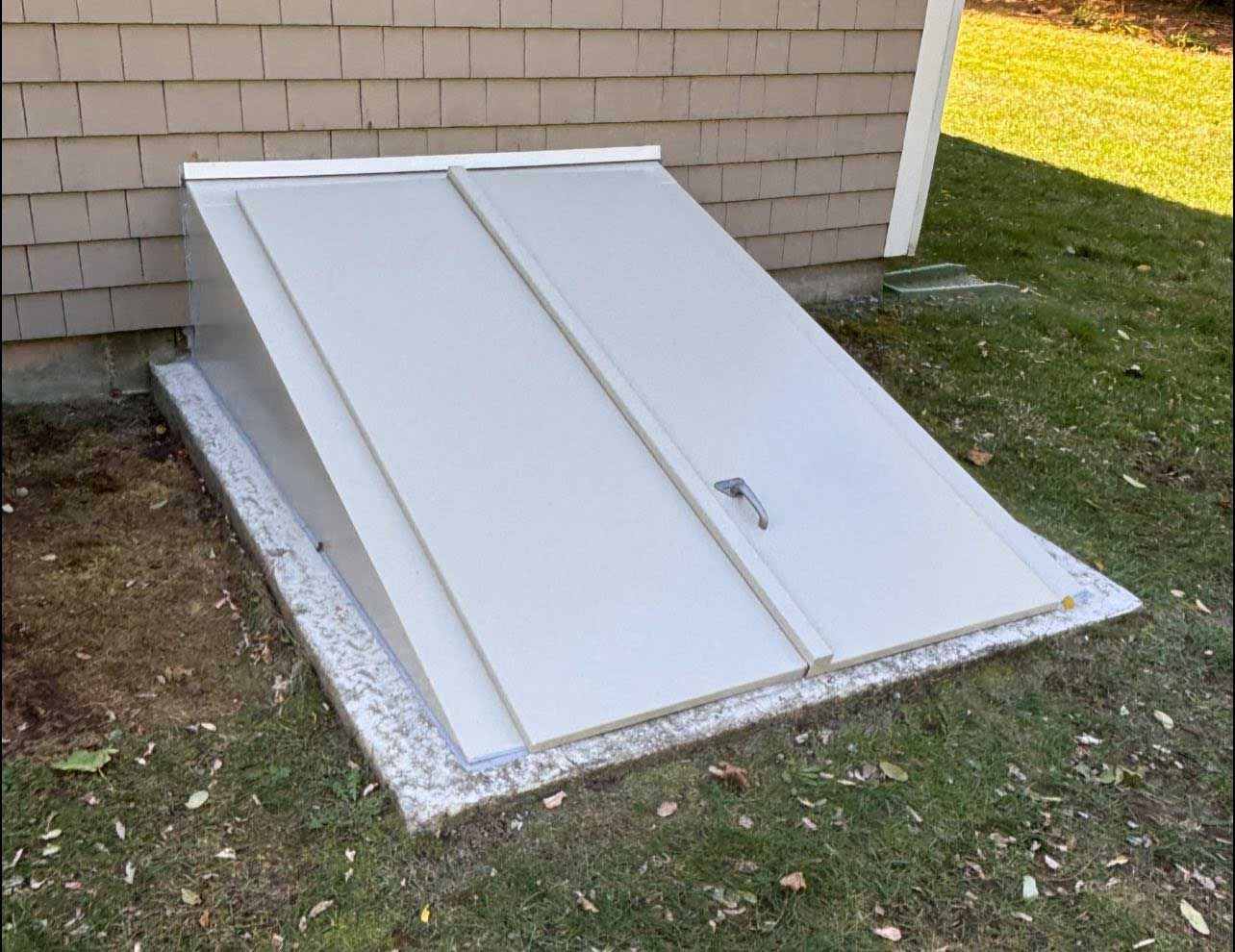 Gray basement hatch with two angled doors on a concrete base, set against a beige house and green grass.