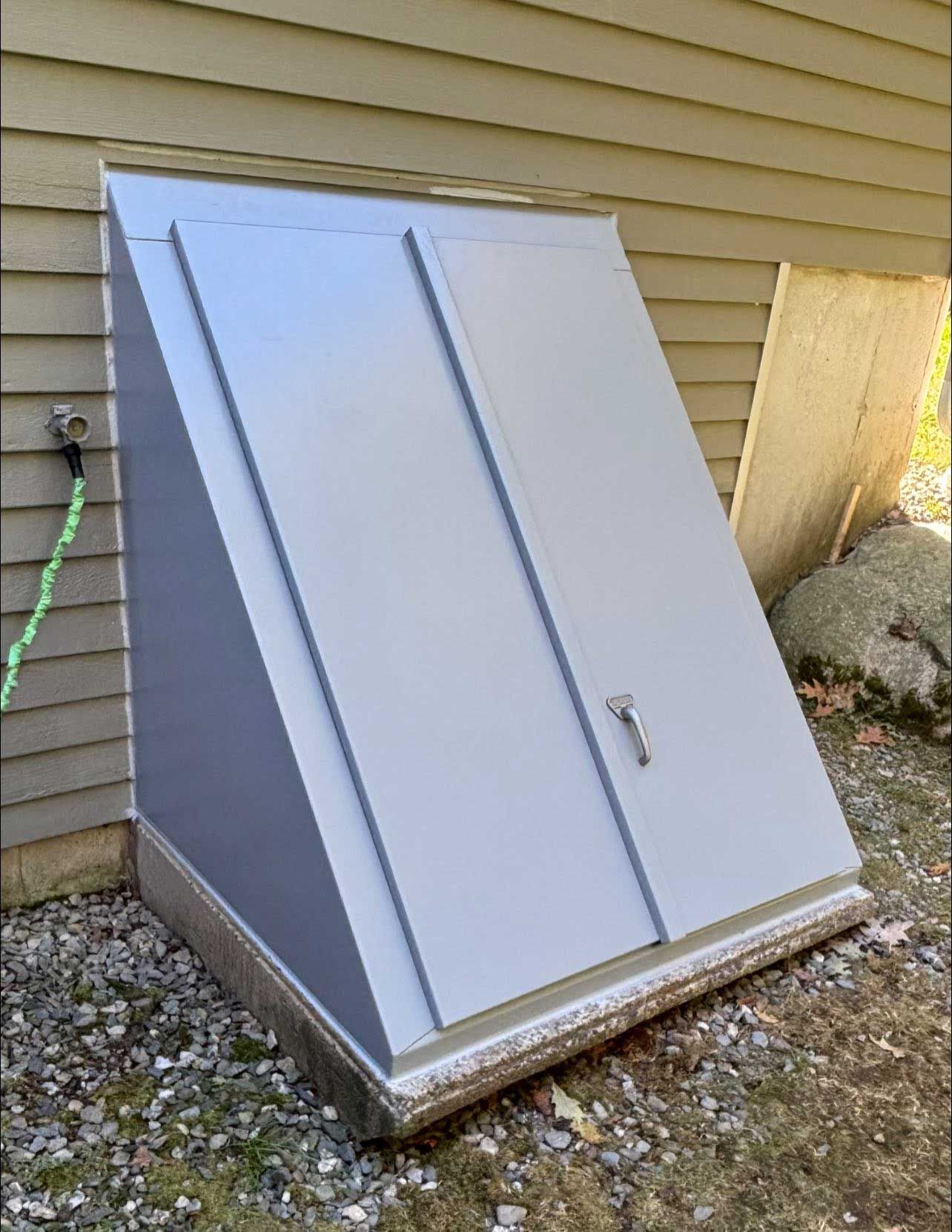 Gray sloped basement door with two panels and handle. Situated on a concrete base next to a tan building.